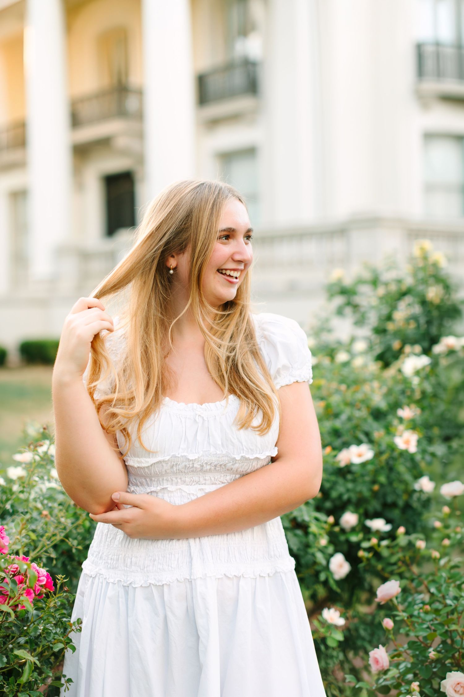 Smiling blonde woman in white dress standing near rose bushes and white columns.