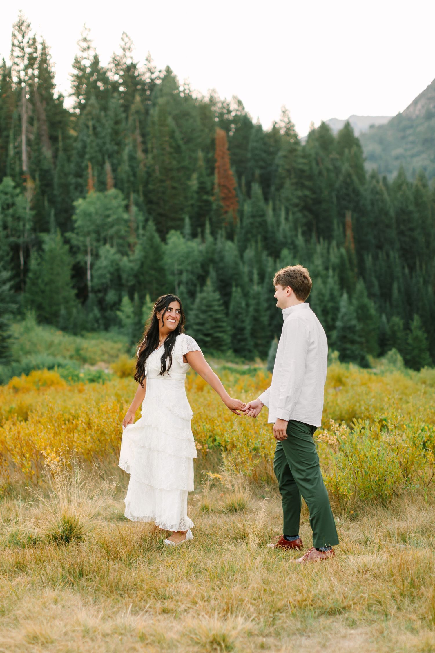 A bride and groom hold hands in a mountain meadow, surrounded by evergreen trees and golden wildflowers.