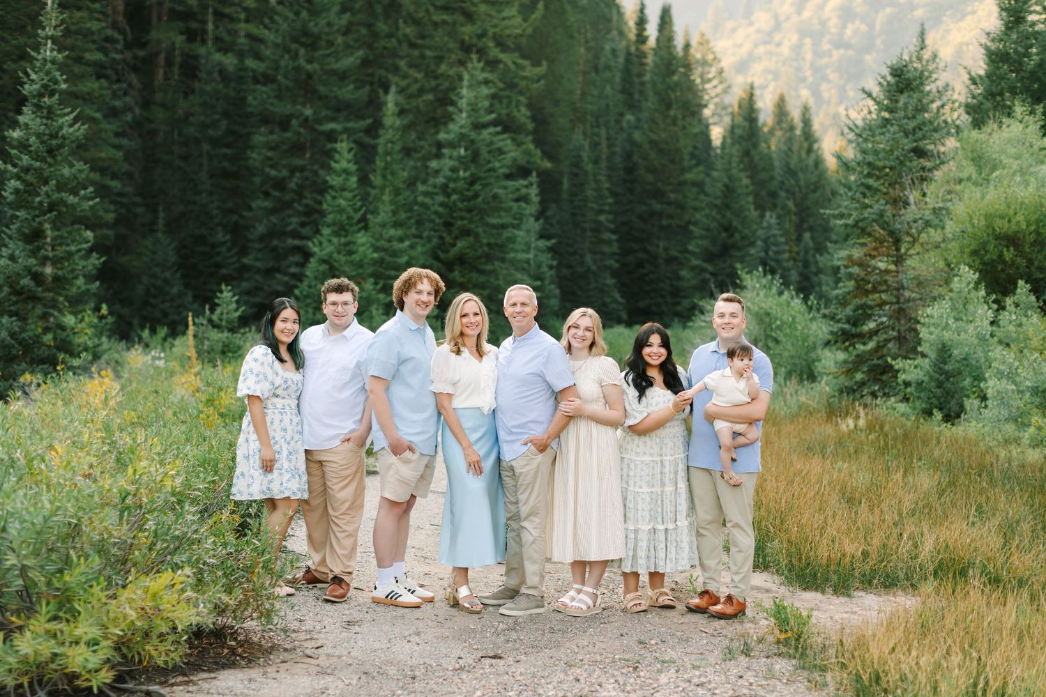 Large family group portrait outdoors in a mountain meadow surrounded by tall pine trees, dressed in coordinated light casual outfits in Utah