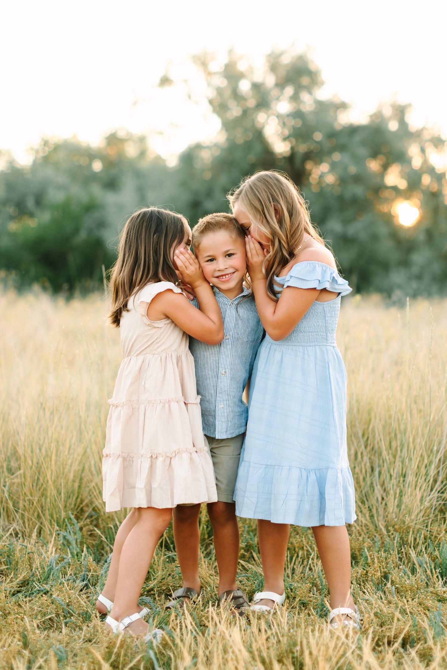 Two girls whispering in the ears of a smiling boy in a sunny field, all dressed in pastel summer outfits in Bountiful, Utah.