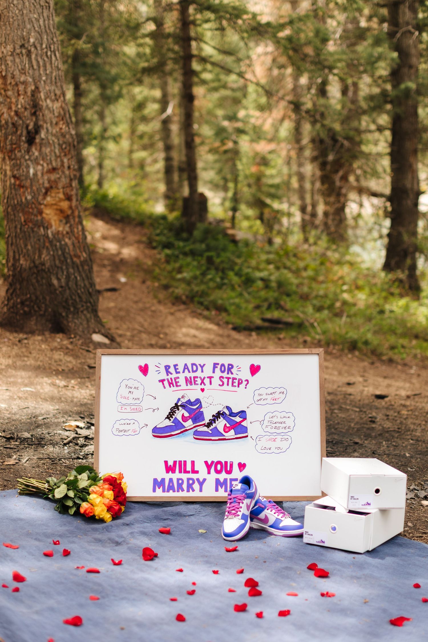 Romantic outdoor marriage proposal setup with sneakers, roses, and a sign reading Will You Marry Me in a forest.