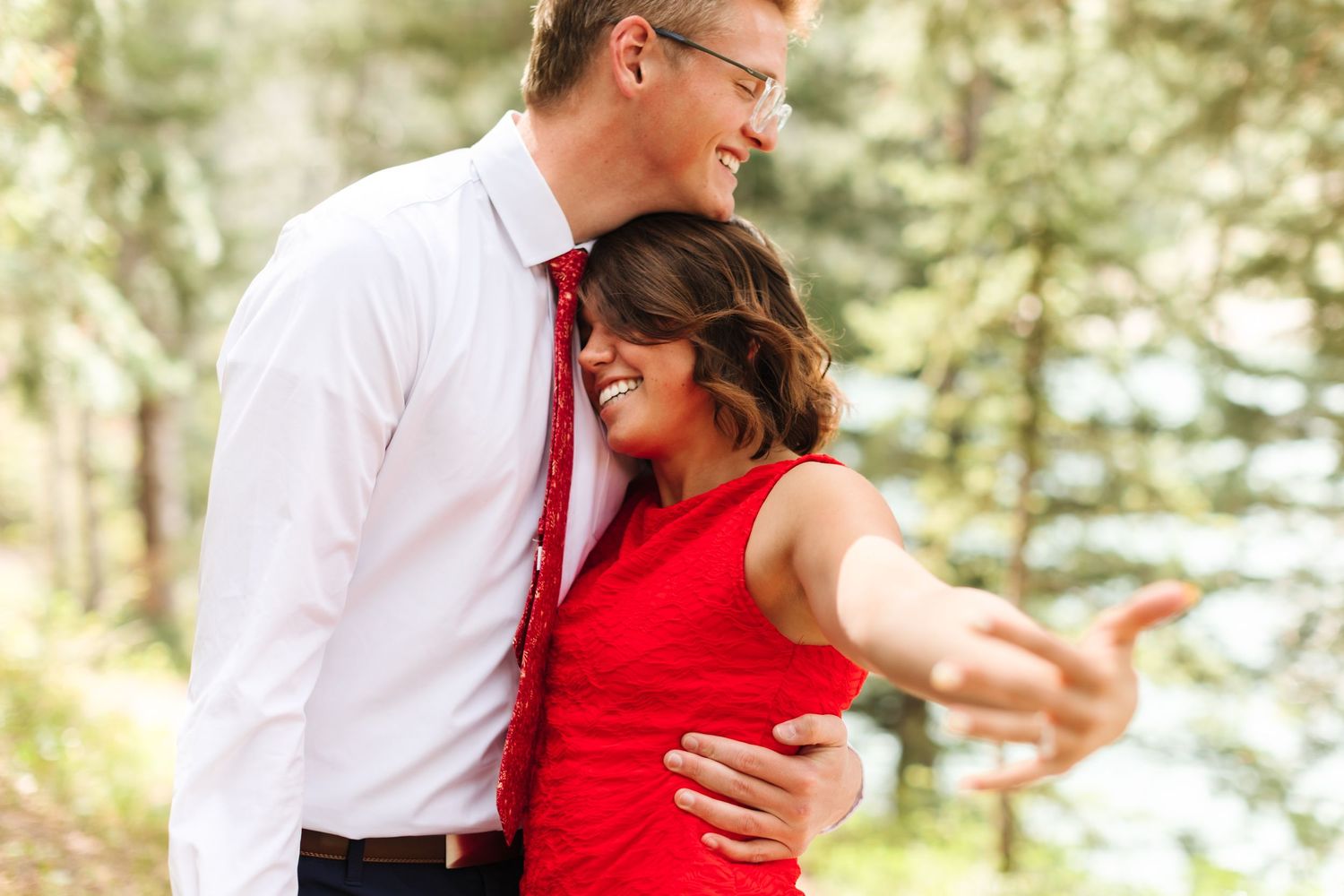 Happy couple embracing outdoors, woman in red dress and man in white shirt with red tie, smiling in a forest setting in Utah.