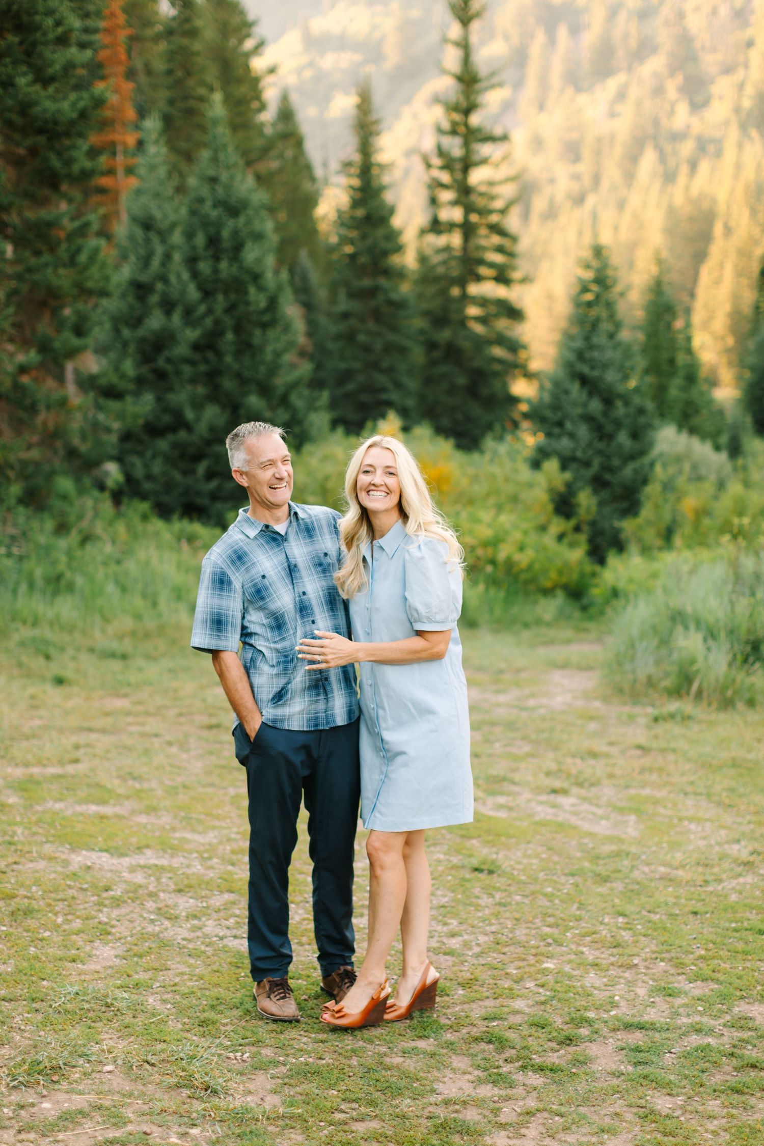 A smiling couple poses together in a lush mountain meadow surrounded by pine trees and golden sunlit hills.