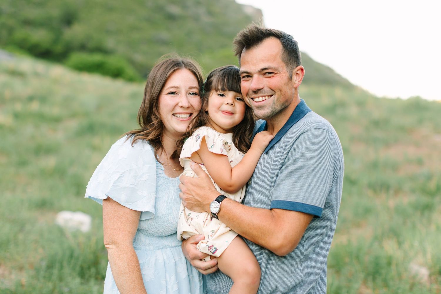 Happy family of three posing outdoors in green hills, parents holding young daughter in floral dress in Provo, Utah.