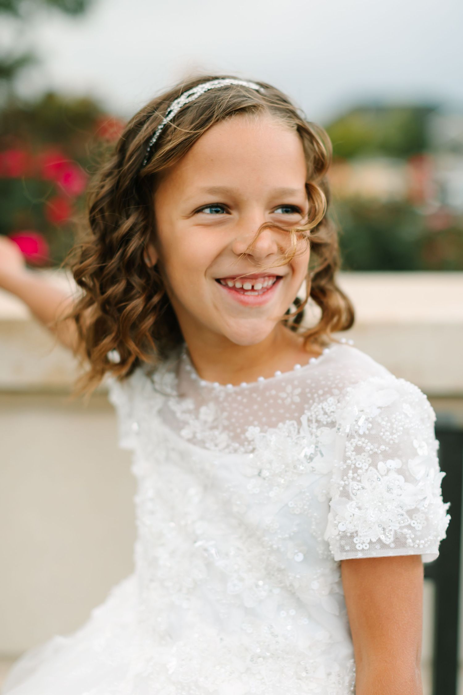 A young child in a white lace dress with delicate details smiles brightly outdoors.