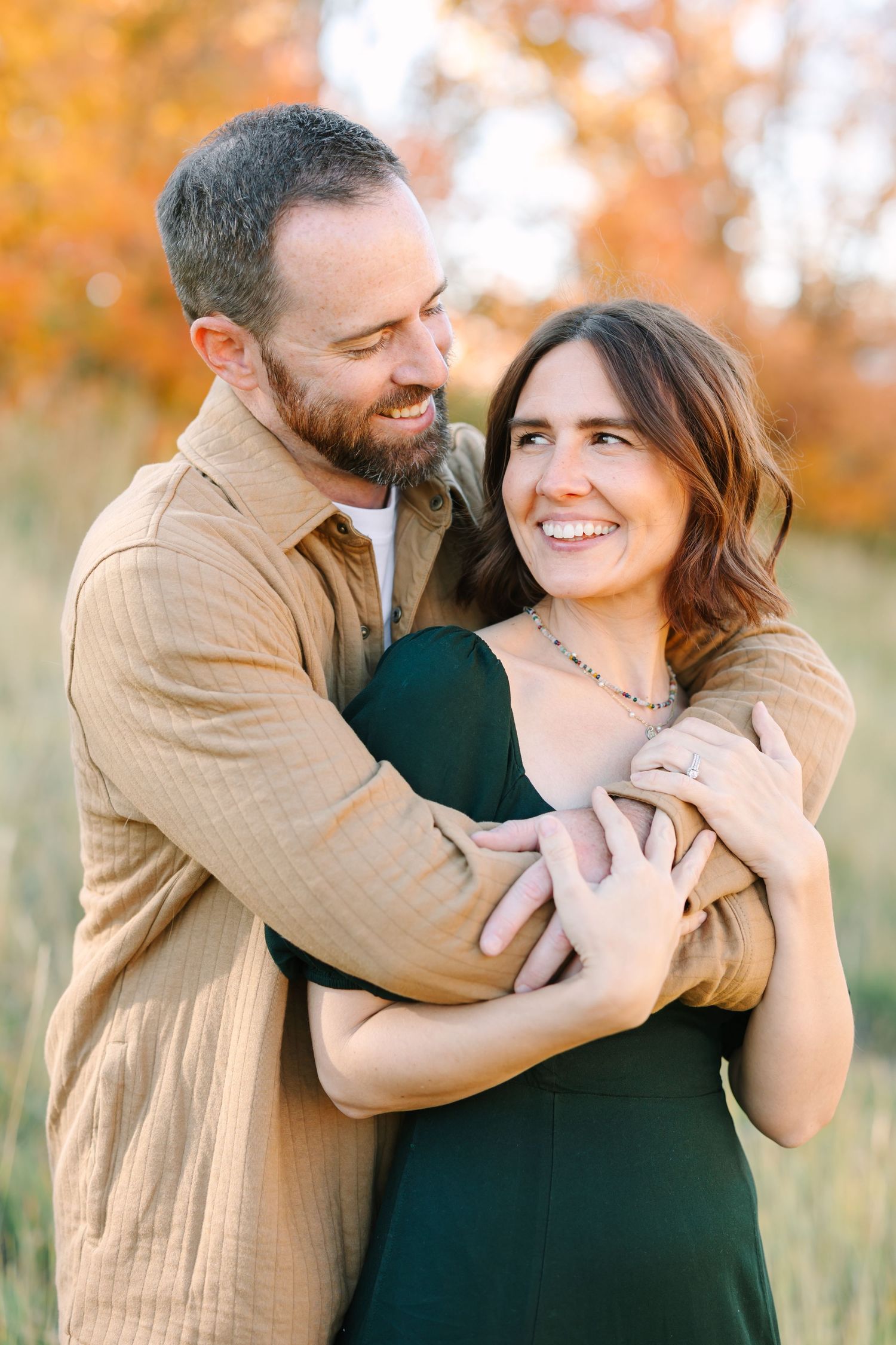A couple embraces and smiles at each other against a backdrop of warm autumn foliage in golden light.