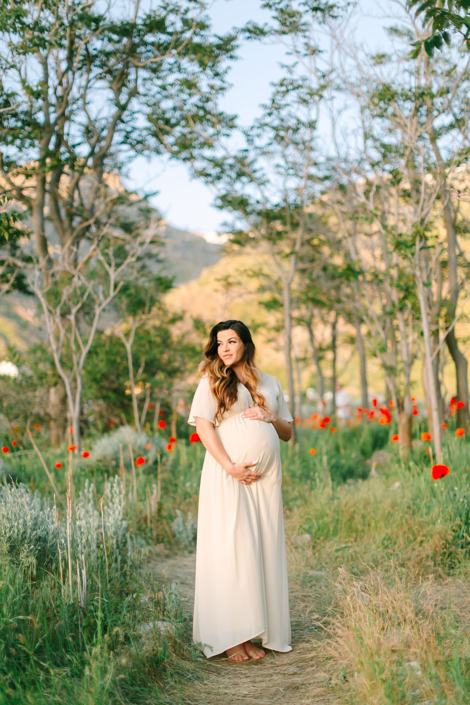 A dreamy maternity photoshoot in a field with tall grass, trees and red poppies at golden hour.