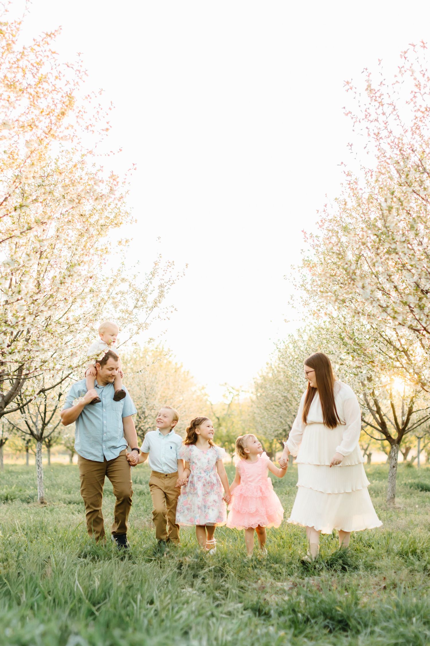 Family of six walking through a blooming spring orchard at sunset, parents with children in pastel dresses and casual attire.