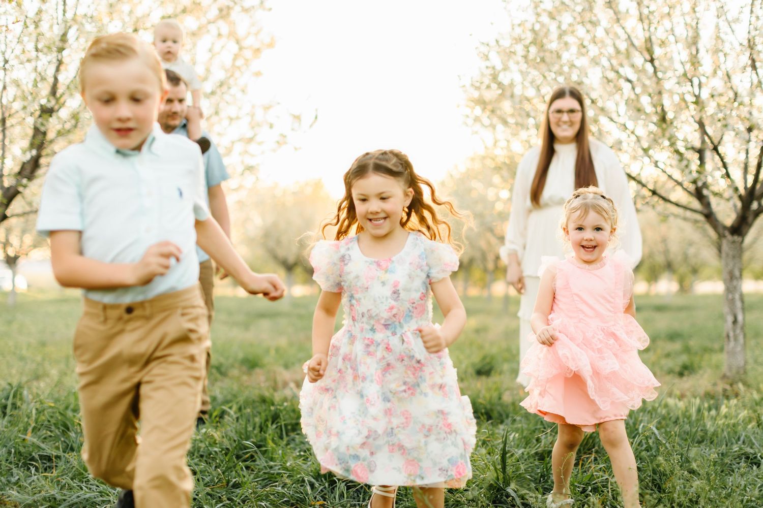 Three children run joyfully through a blossoming orchard at sunset while two adults watch warmly in the background.