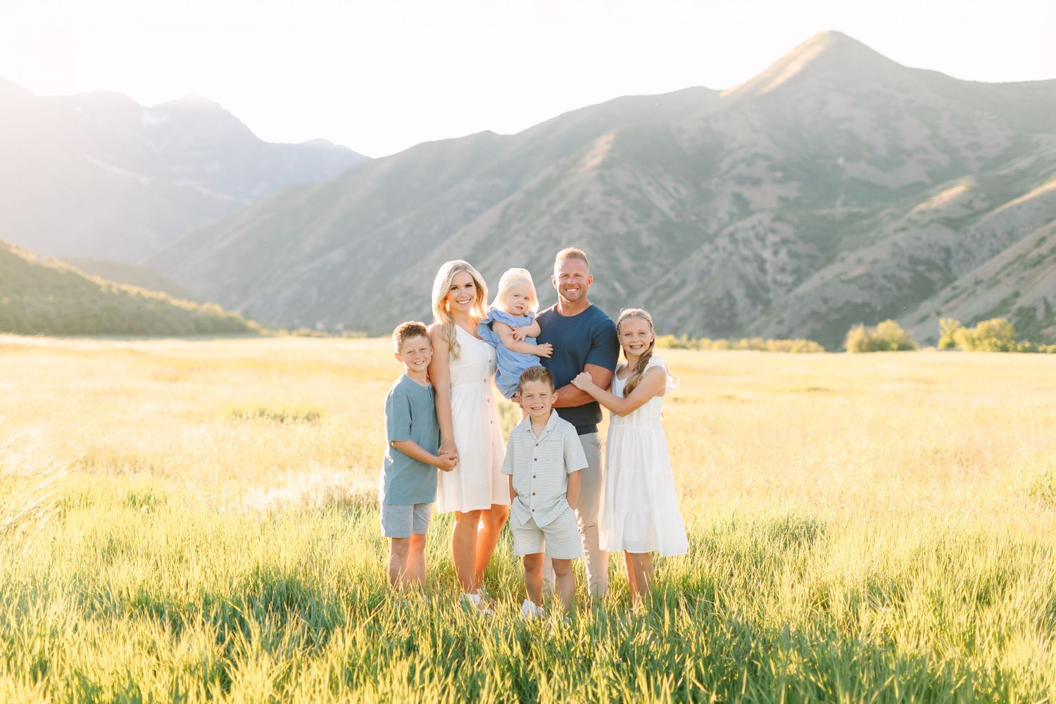 Family of seven posing in a sunlit mountain meadow, wearing coordinated white and blue outfits for a professional outdoor portrait.
