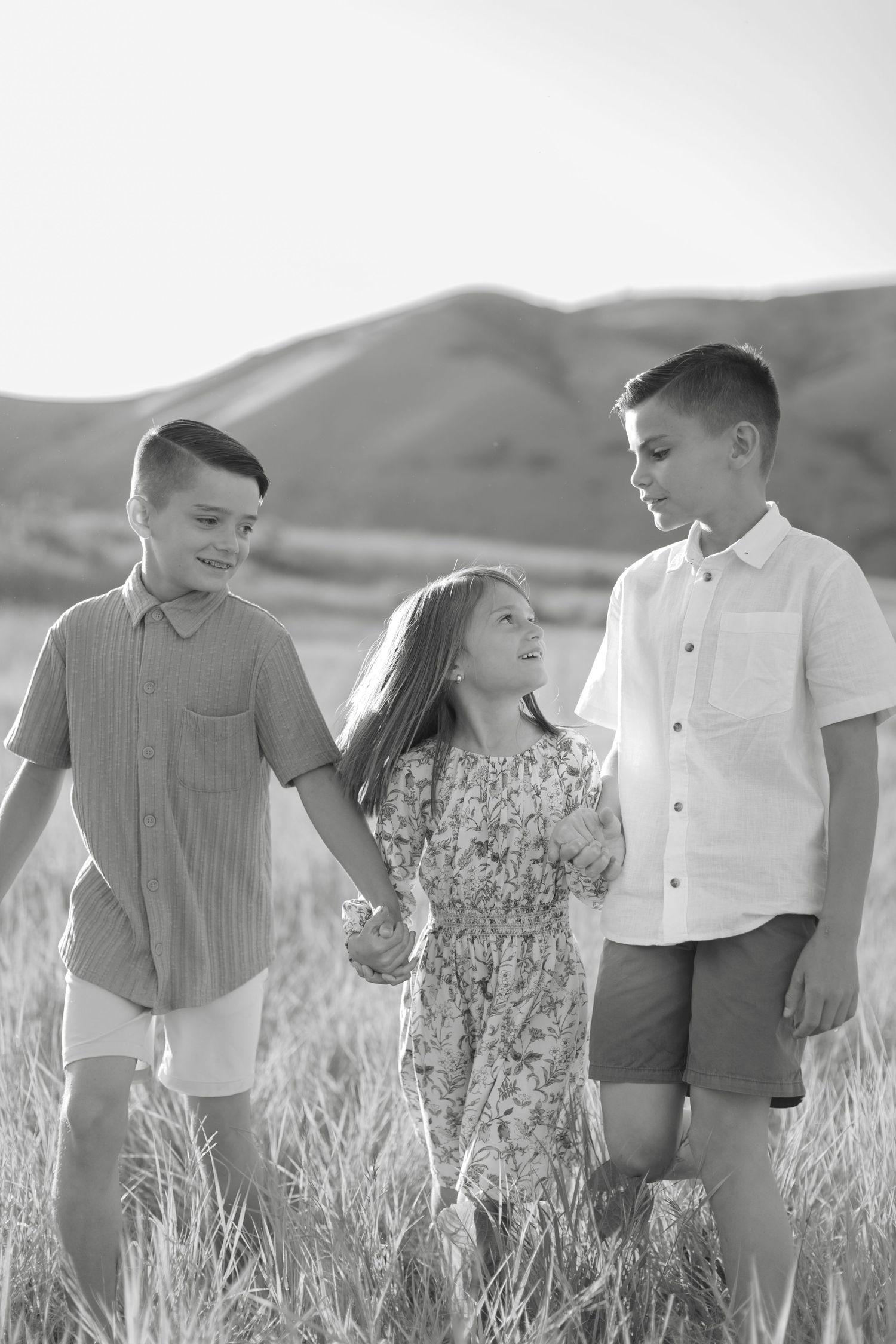 Three children stand together in a grassy field with rolling hills in the background in black and white.