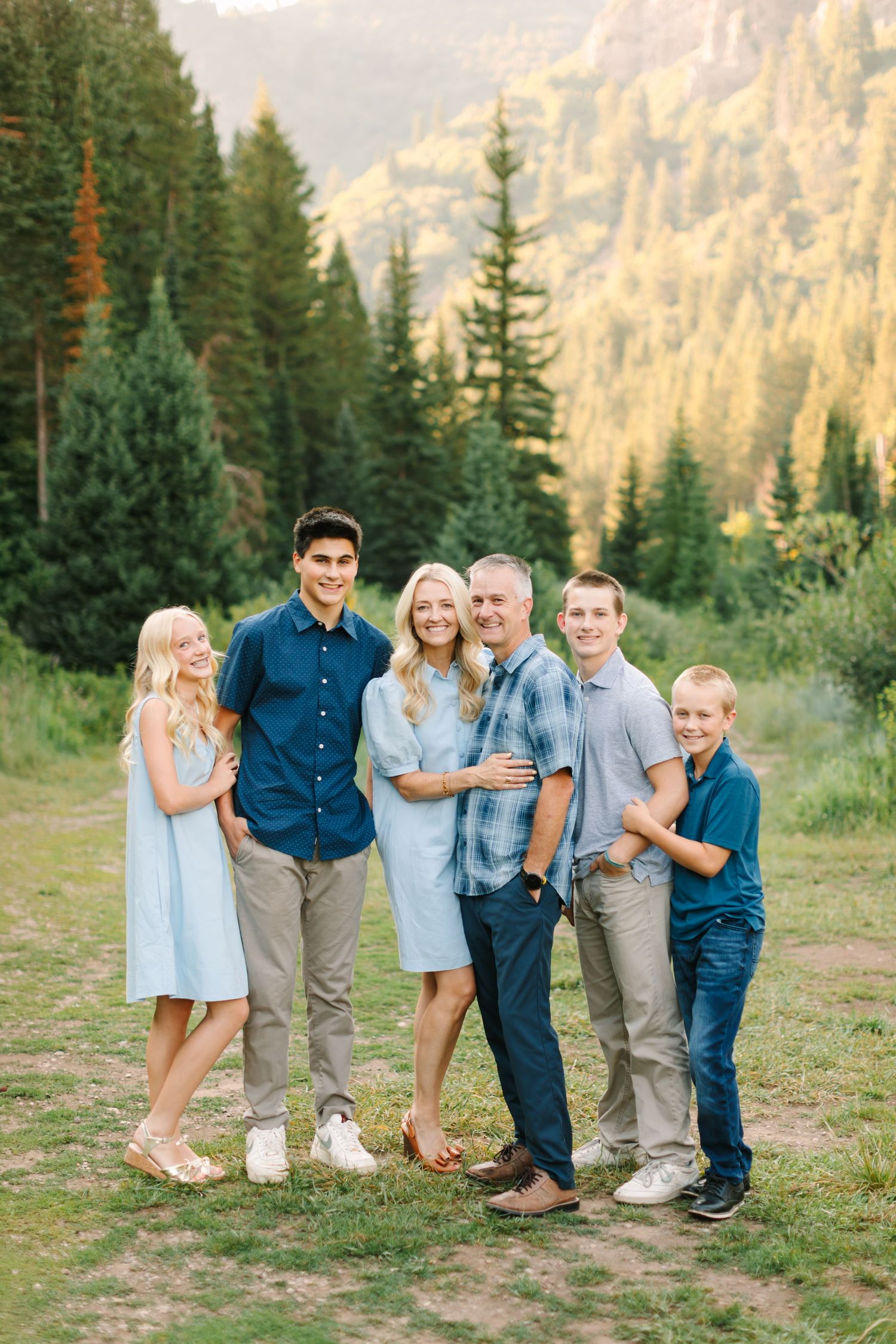 Family of six posing outdoors in coordinated blue outfits with mountain forest backdrop during golden hour.