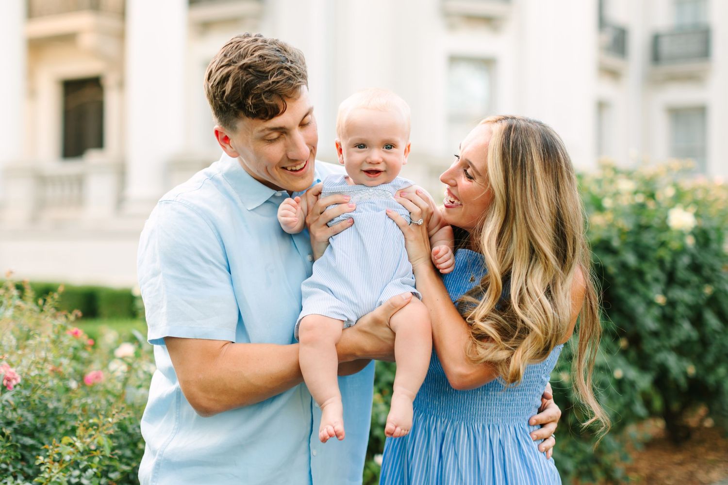 A happy young family in coordinating blue outfits holds their smiling baby outdoors near blooming roses.