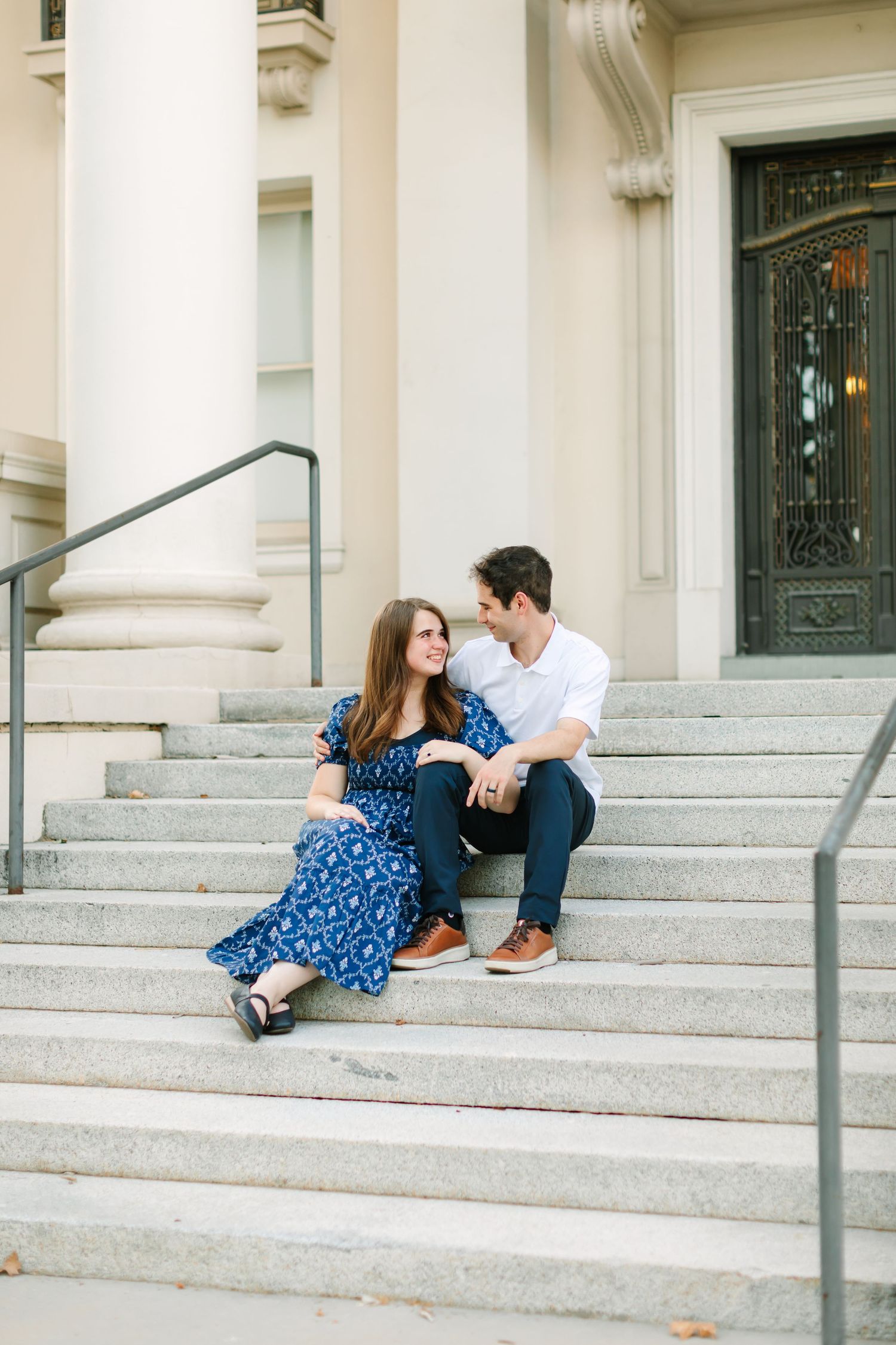 Couple sitting on stone steps of a classic building, woman in blue floral dress smiling at man in white shirt and navy pants.
