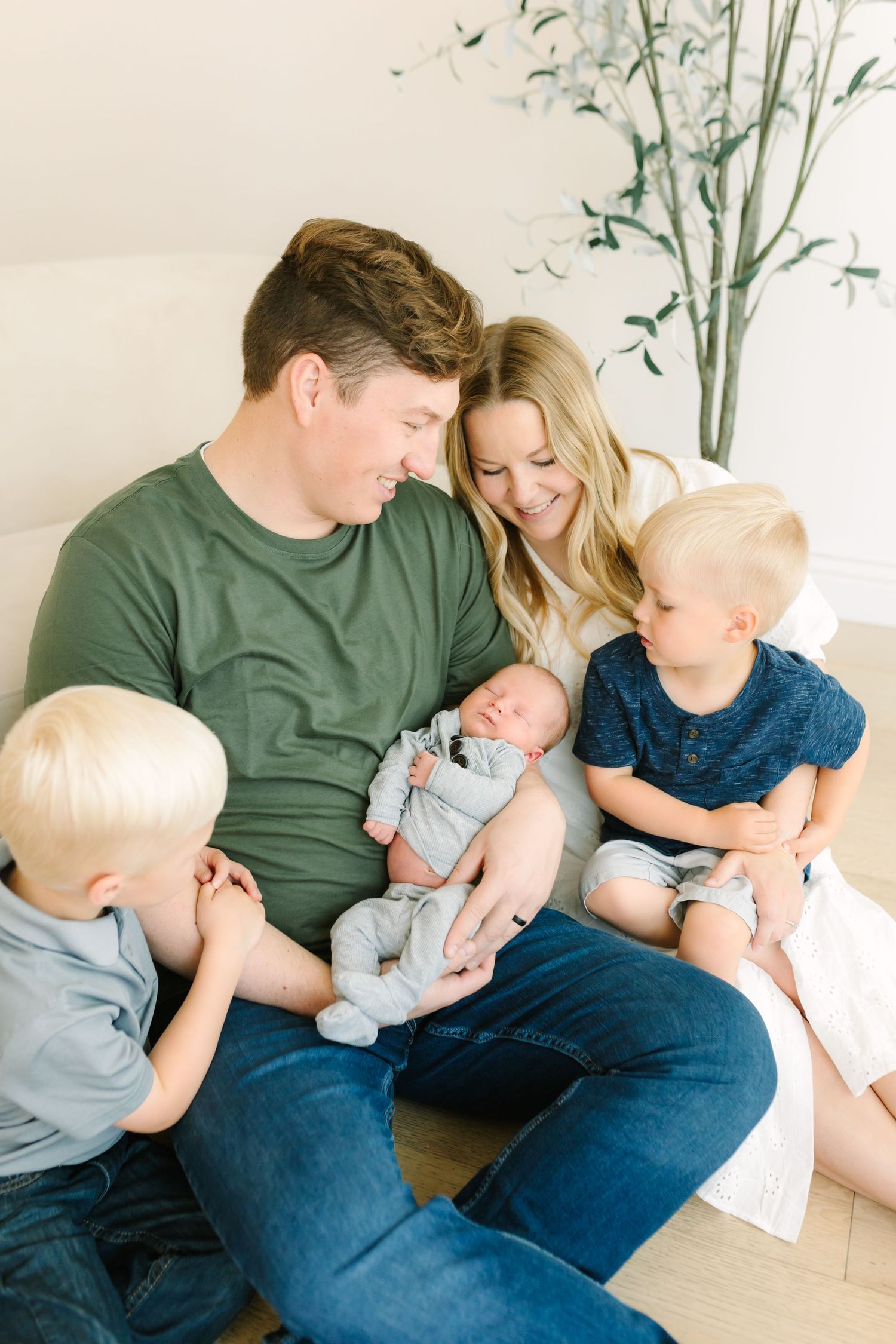 A happy family of five sitting together, parents gazing at their newborn baby while two young boys look on.
