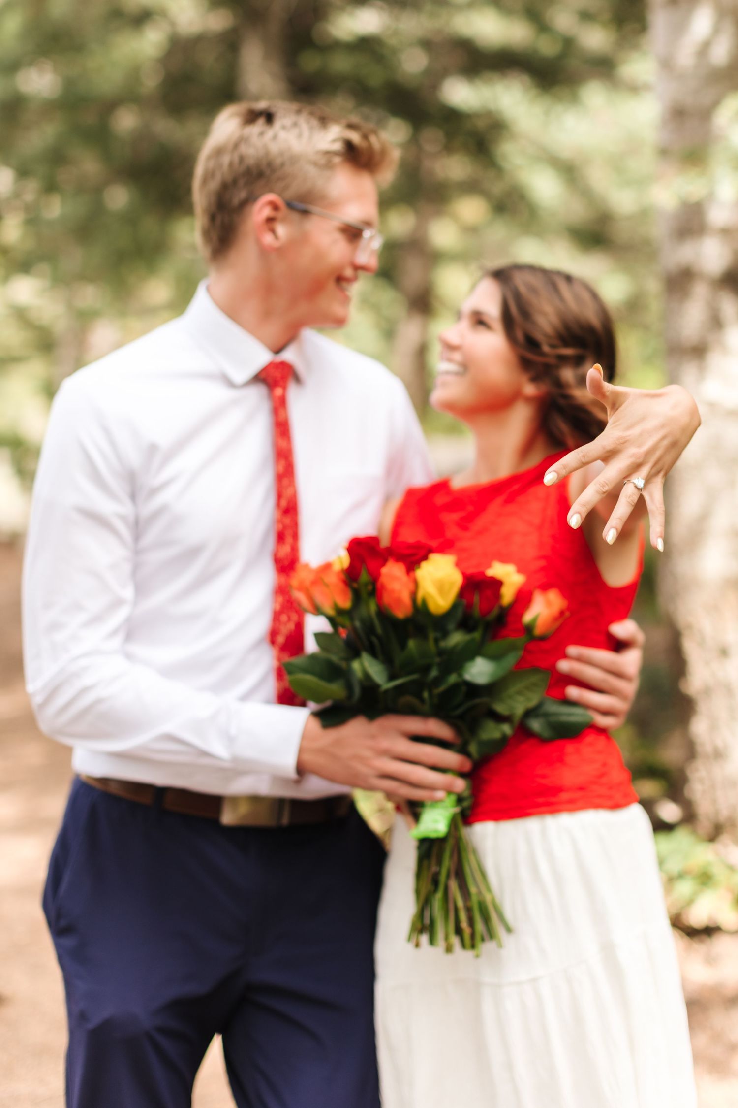 Smiling couple in forest, man in white shirt and red tie holding colorful roses, woman showing engagement ring.