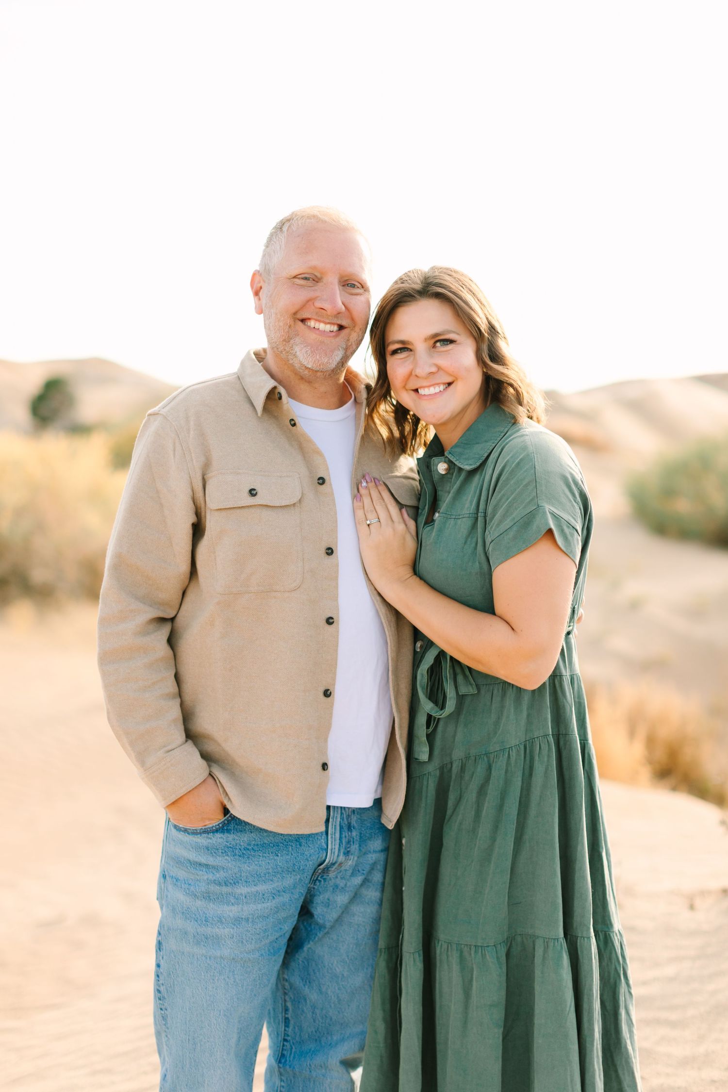 A couple embraces while standing in a desert landscape during golden hour wearing casual clothing in green and tan.