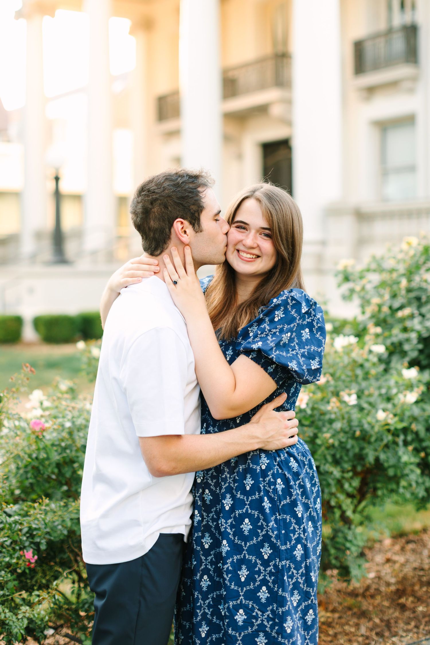 Couple holding hands smiling at each other in front of a grand white neoclassical building with columns and rose bushes in Utah.