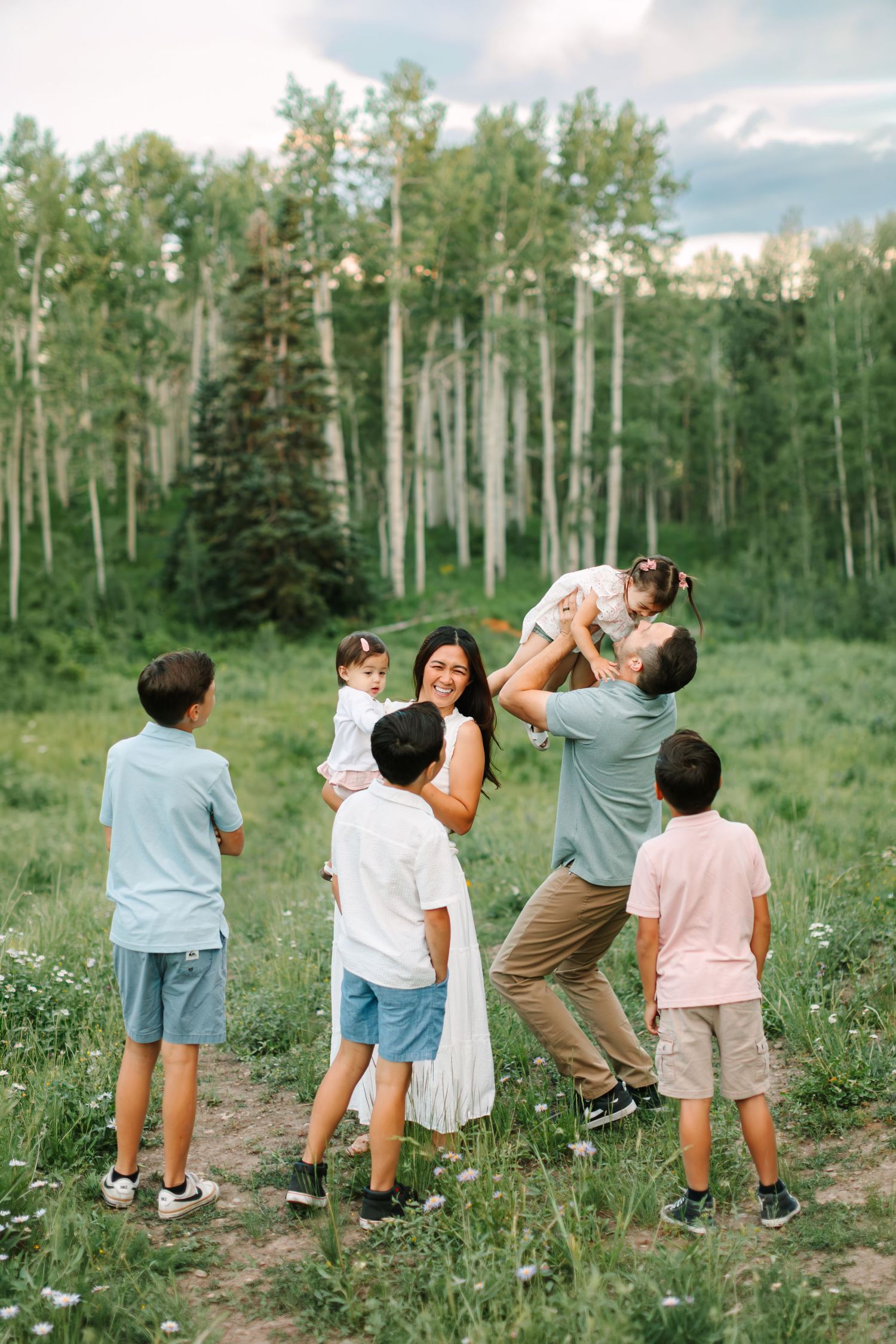 Family of six enjoying a playful outdoor moment in a lush green forest clearing, father lifting daughter joyfully in Heber, Utah.