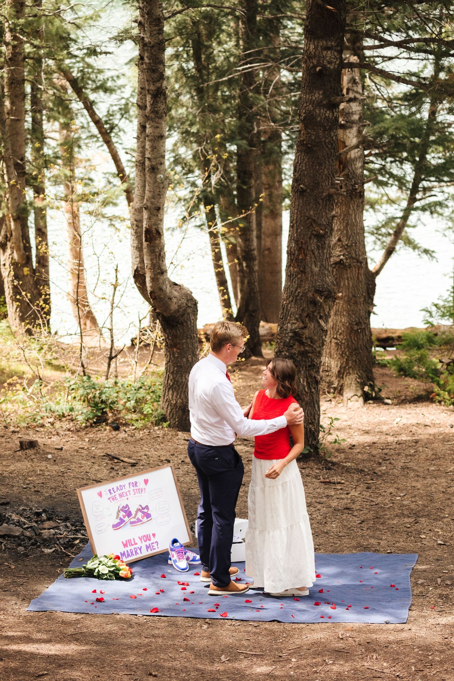 Couple sharing a romantic proposal moment in a forest clearing with a sign and flower petals on a blanket.