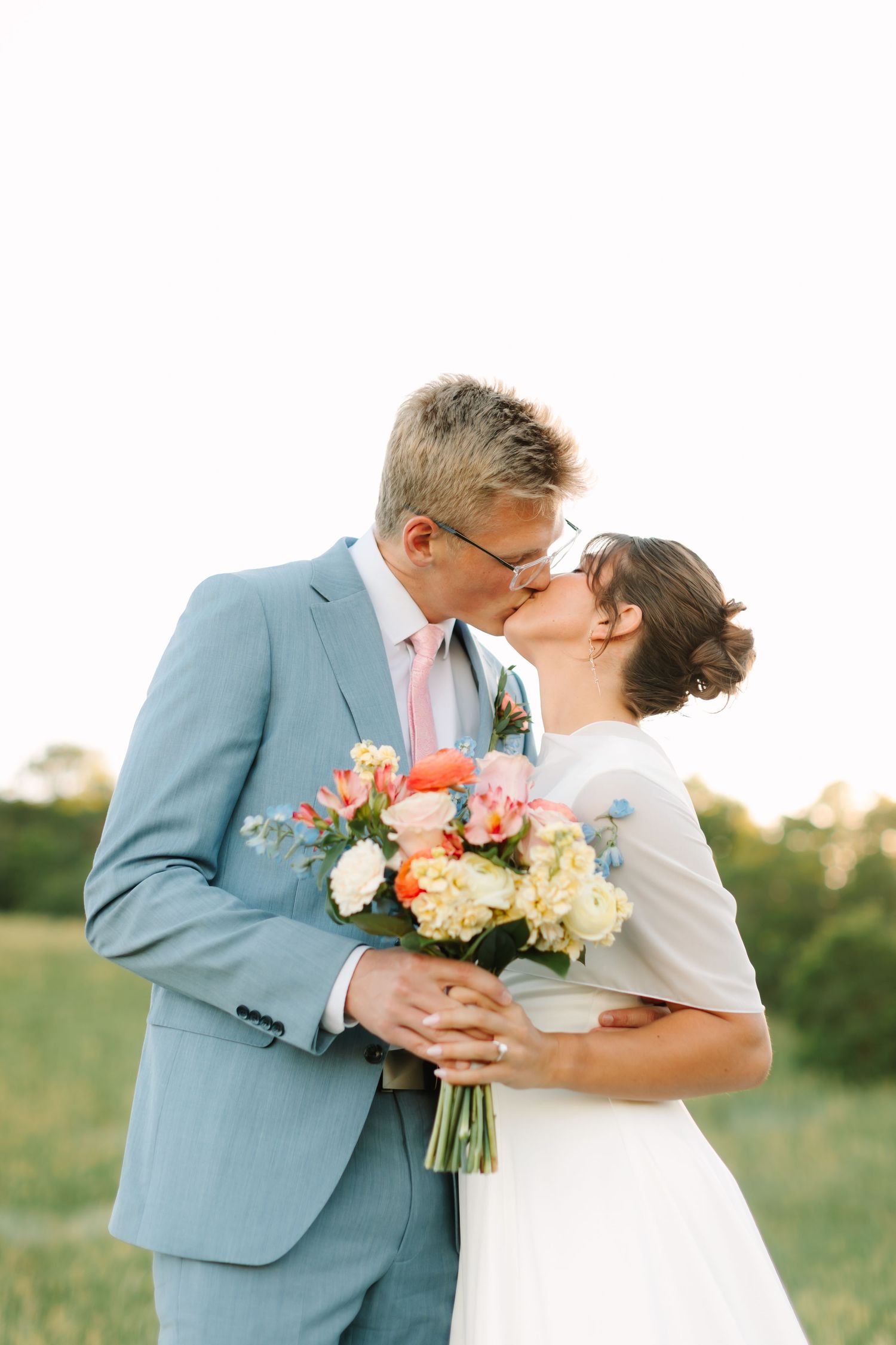 Bride and groom share a romantic kiss outdoors, holding a colorful bouquet of pink and white flowers on their wedding day.