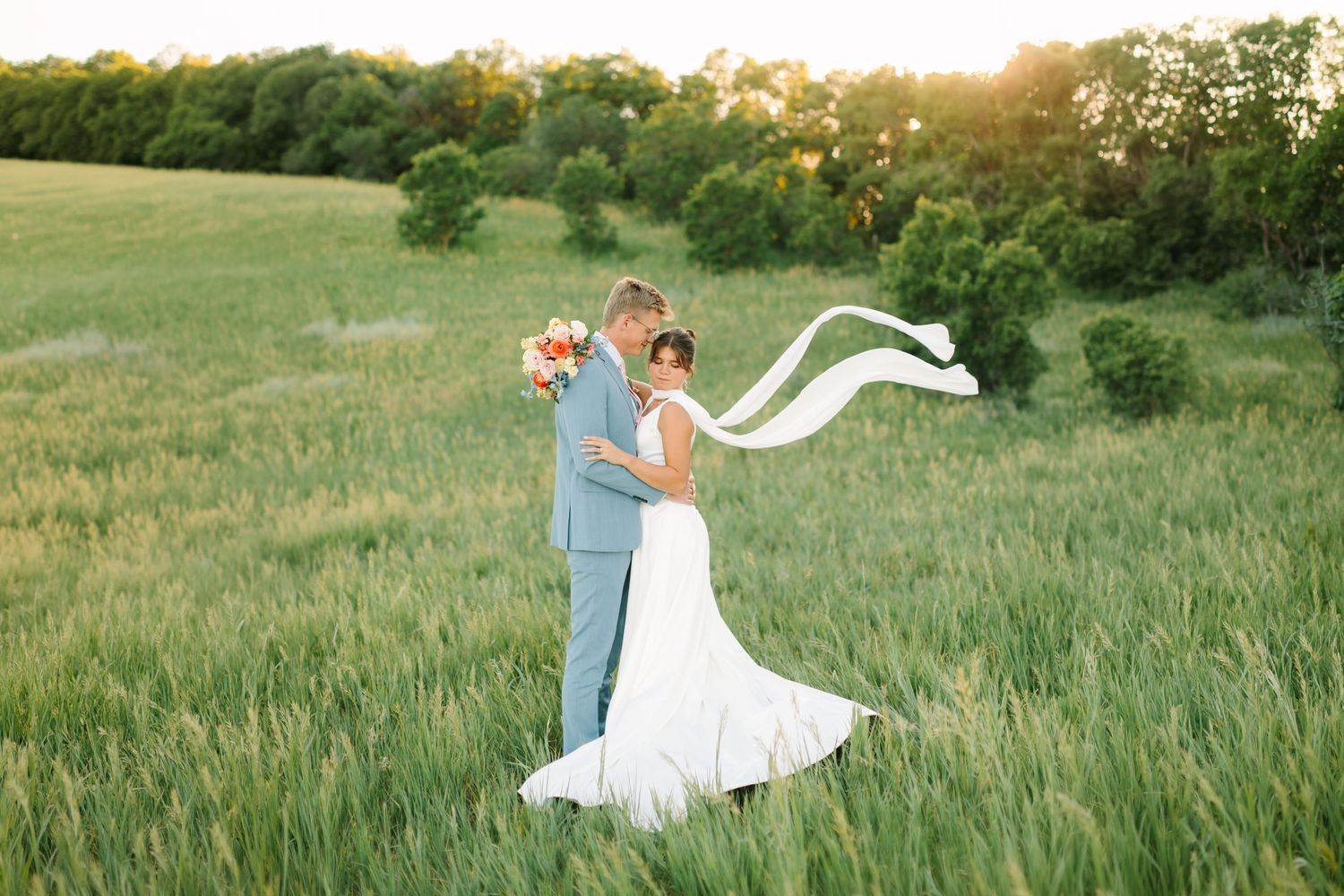 Bride and groom embrace in a sunlit green field, her white gown and ribbon flowing in the breeze.