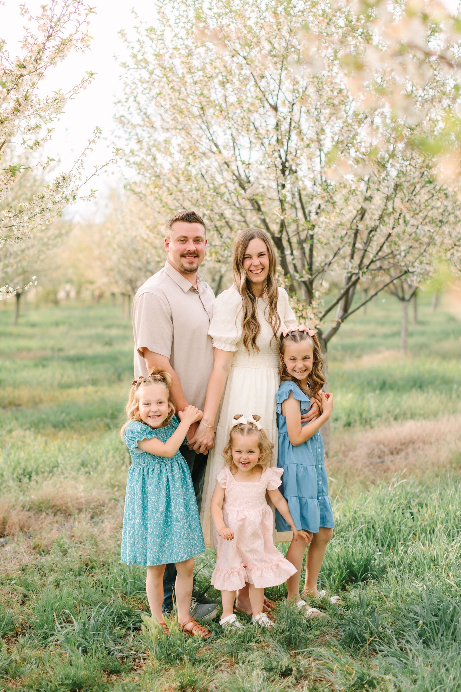 Family of five posing in blooming orchard, parents with three young daughters in colorful spring dresses.