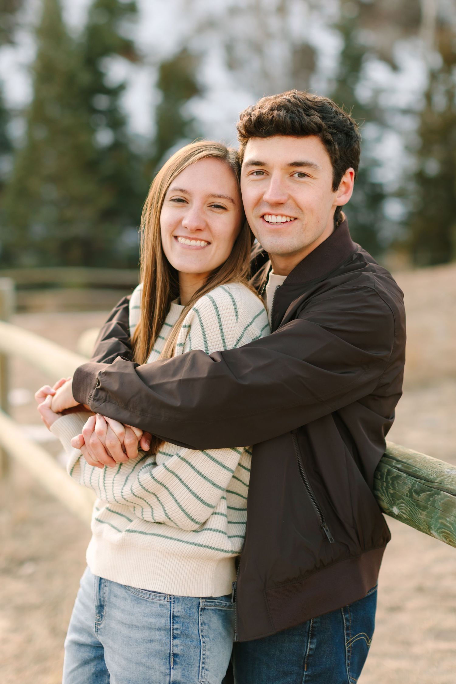 A couple embraces outdoors on a sunny day, wearing casual clothing with trees in the background.