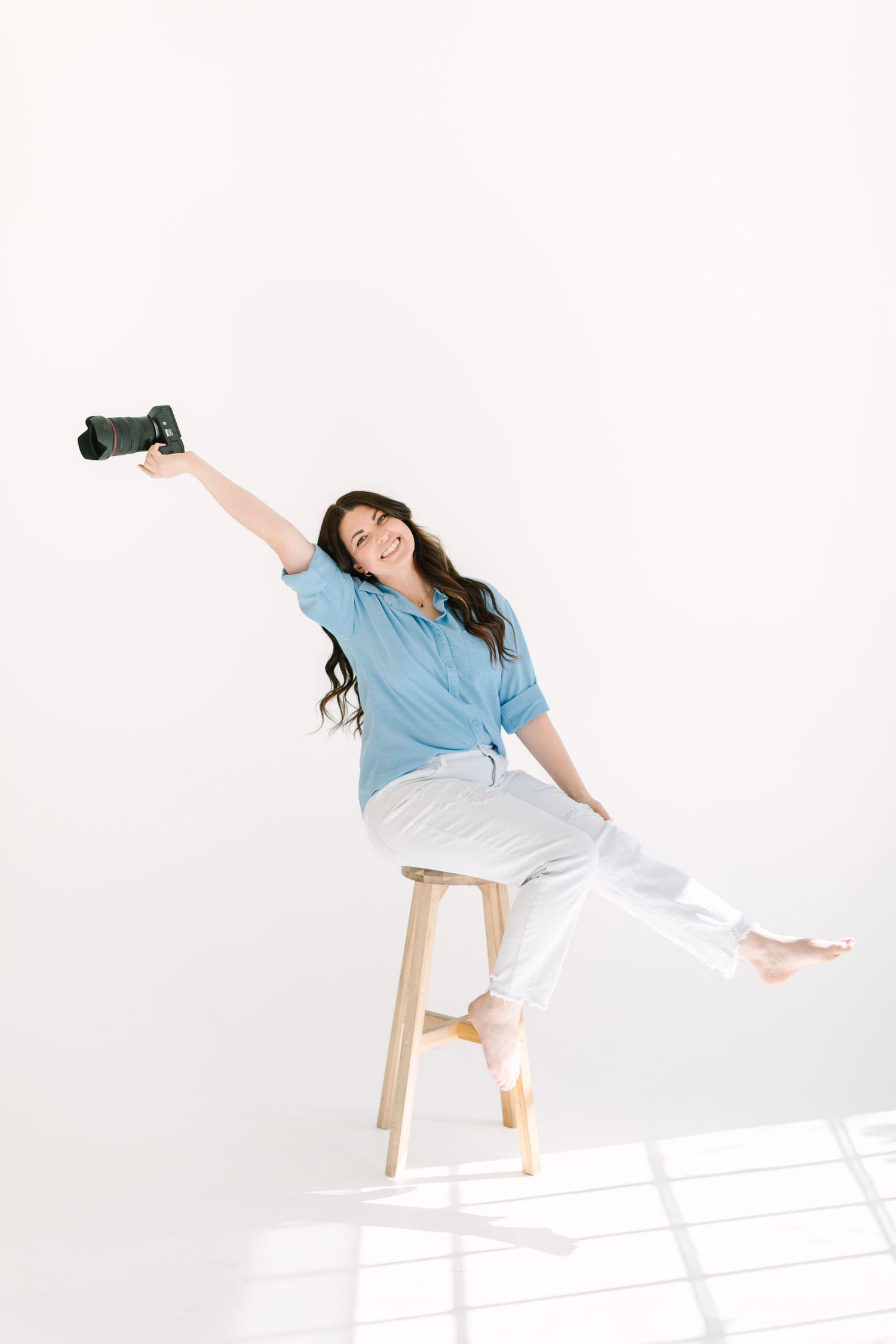 Person in casual attire posing playfully on wooden stool against white backdrop with camera.