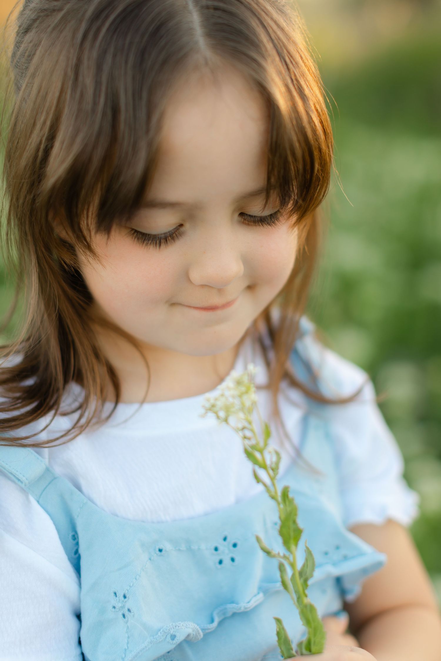A child in a light blue dress smiles gently while looking down outdoors in soft natural lighting.