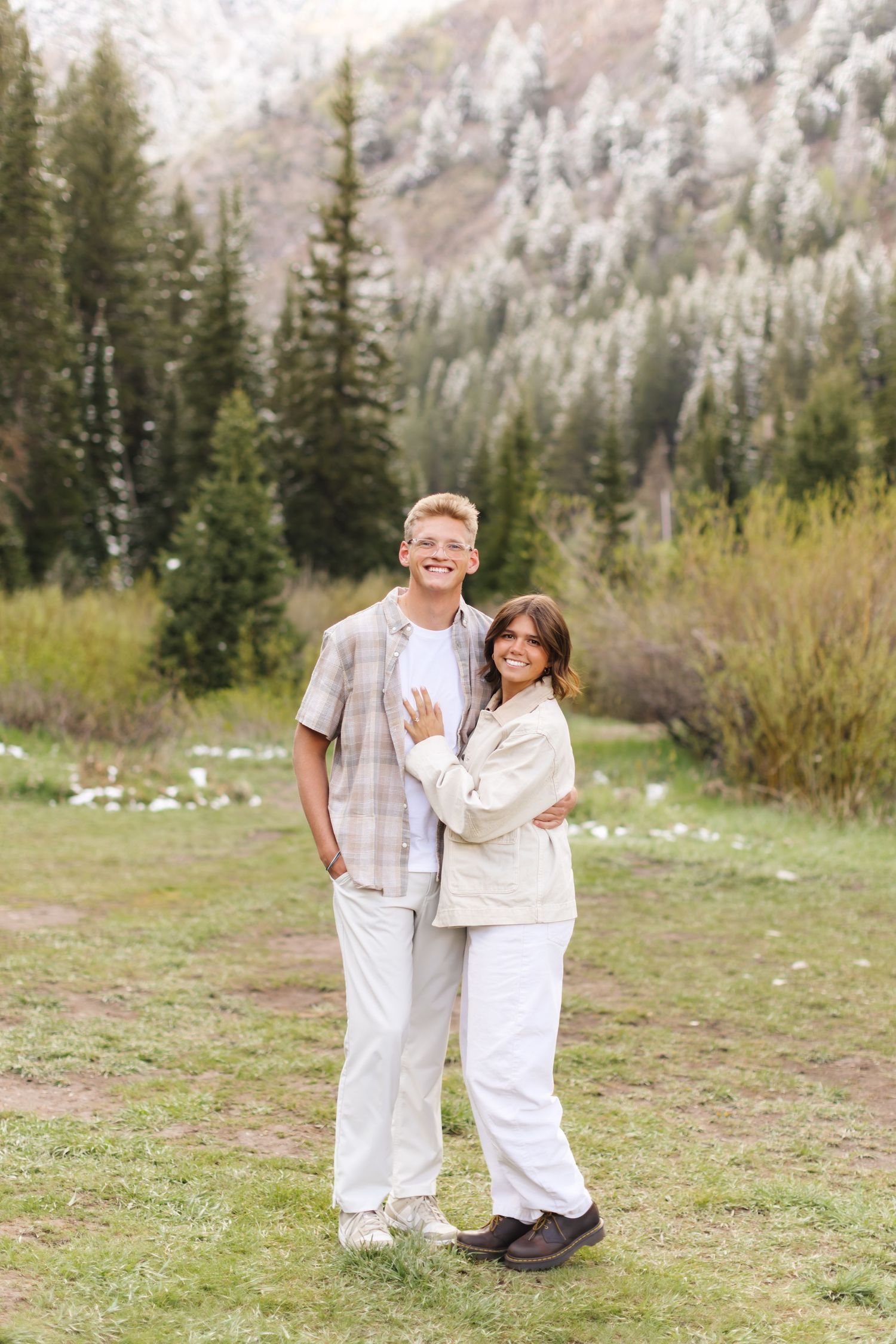 A young couple embracing outdoors in a mountain meadow surrounded by evergreen trees and snow-dusted peaks.