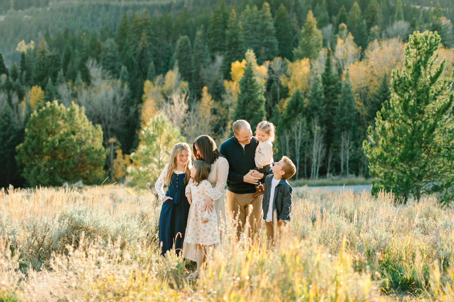 Family poses together in golden autumn field with mountains and evergreen forest in background.