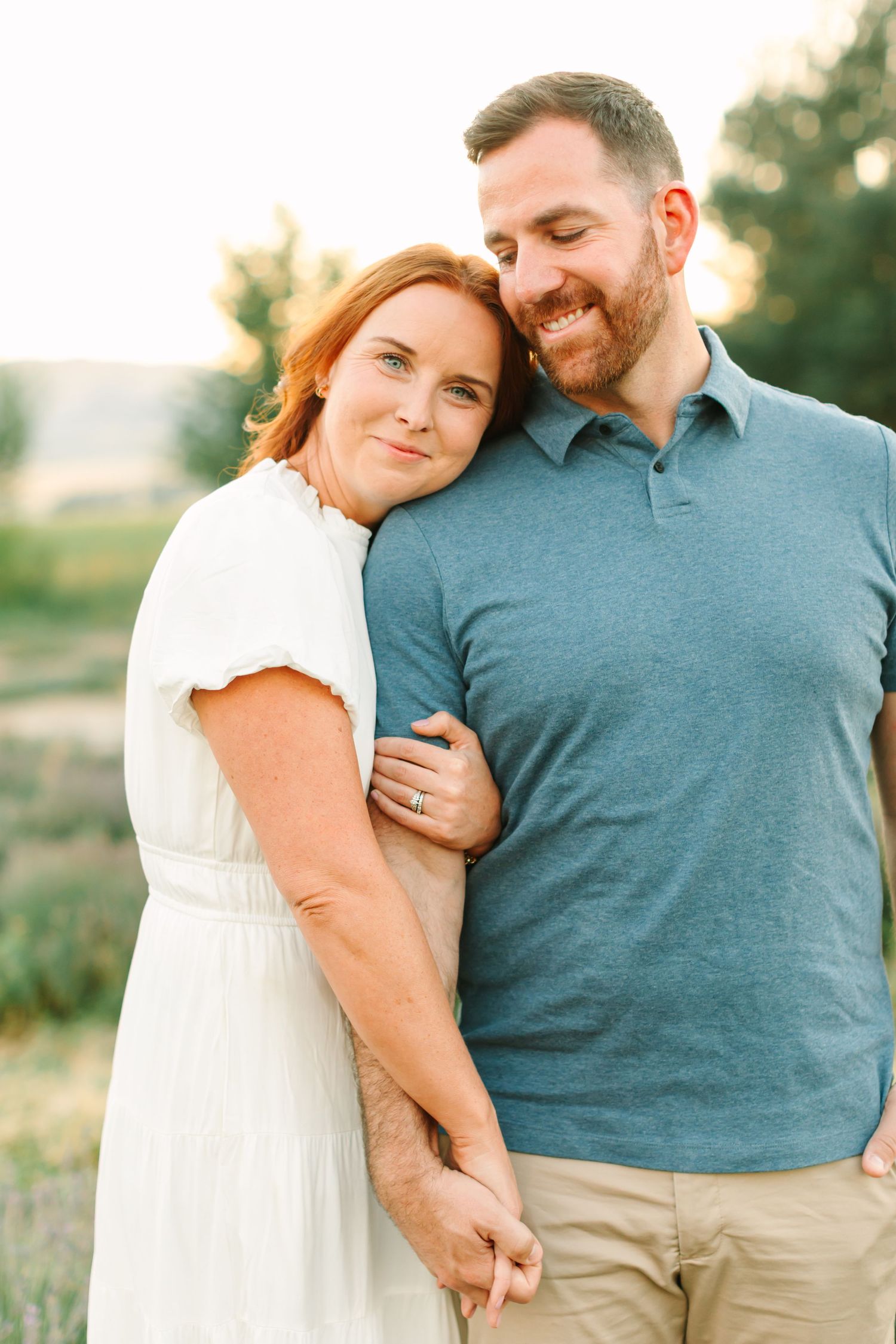 A couple embraces in a romantic outdoor setting during golden hour, wearing white and blue casual attire.