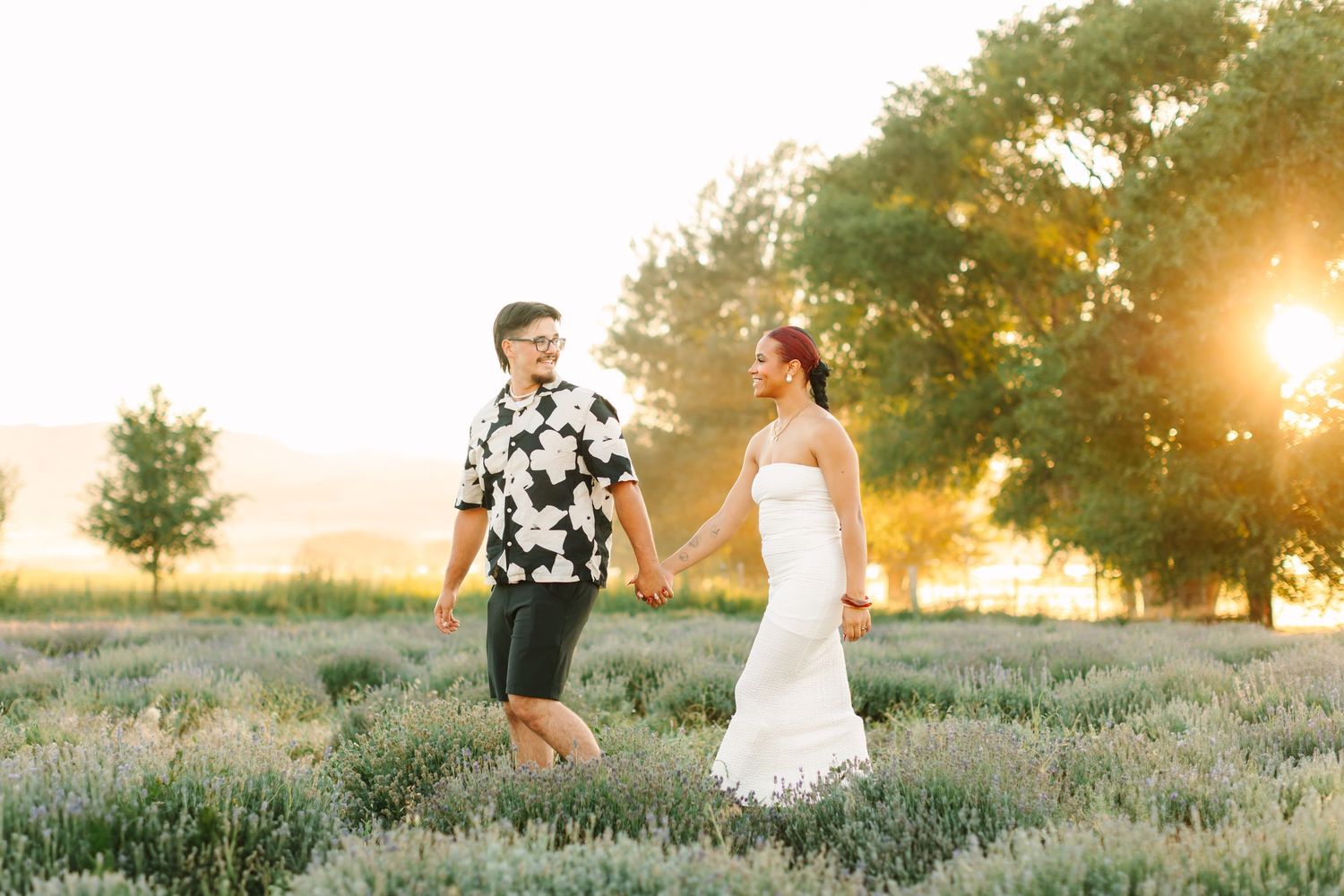 A couple walks hand in hand through a lavender field at sunset while golden light filters through the trees.