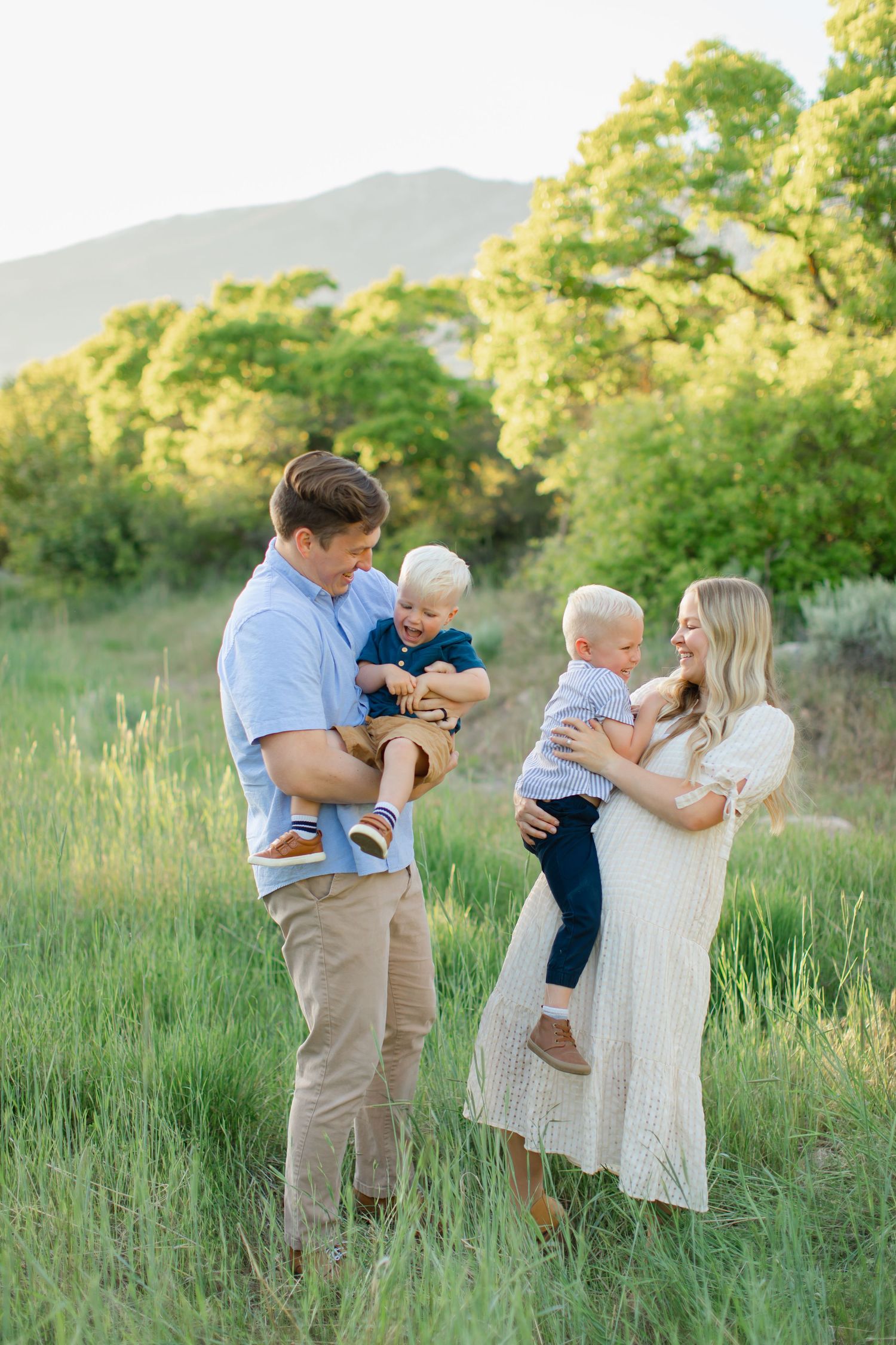 A family shares joyful moments together in a sunlit meadow with tall grass and mountains in the background.