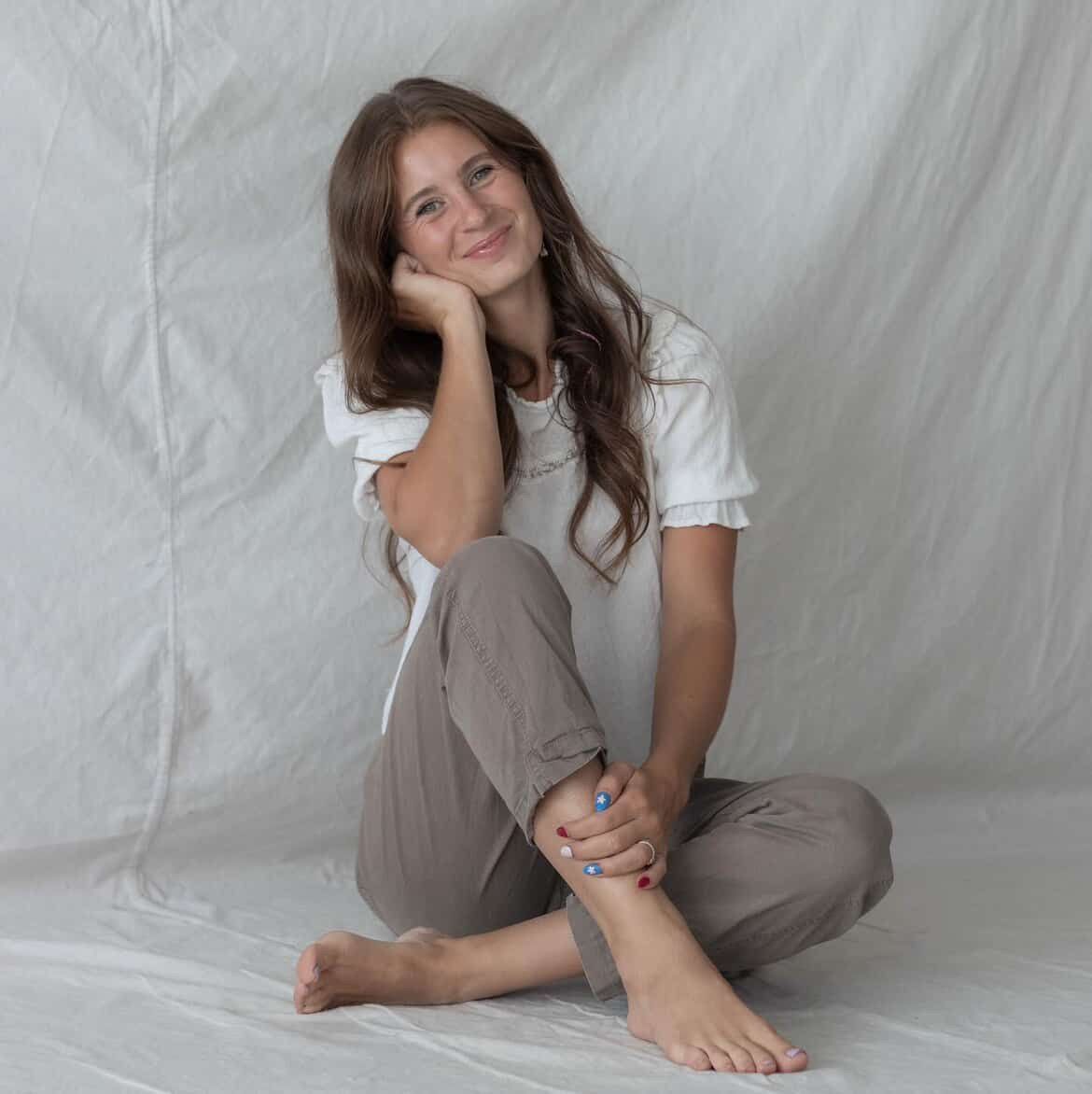 Person in casual white top and beige pants poses playfully against white backdrop in lifestyle photoshoot series.