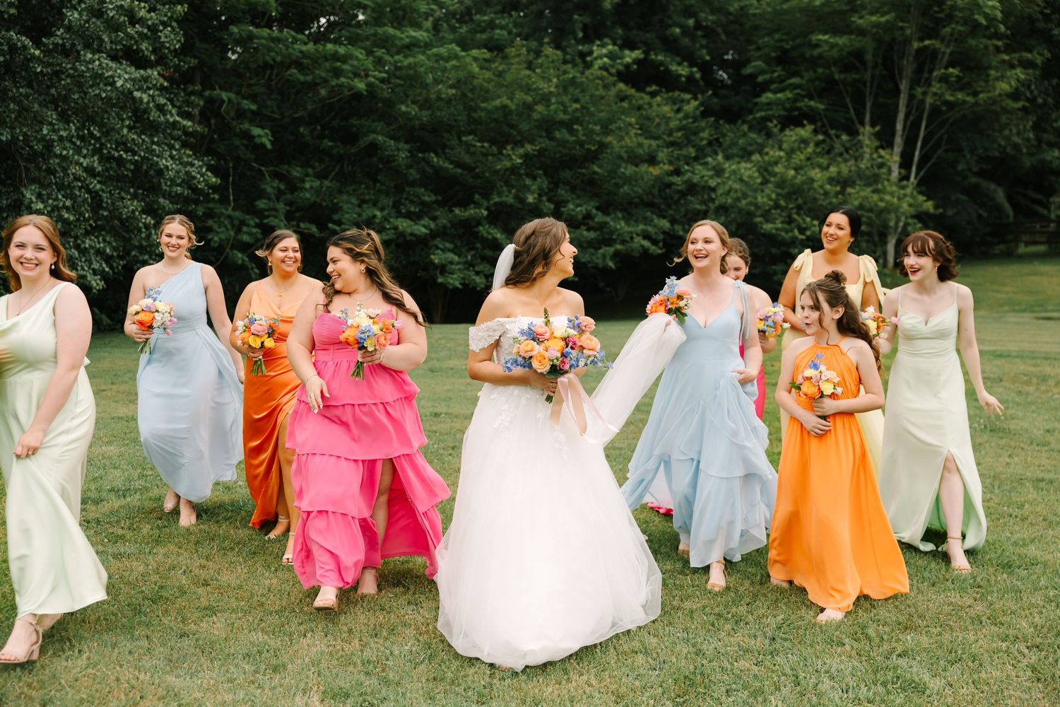 A joyful group in colorful formal attire runs together across a grassy lawn on a summer day.
