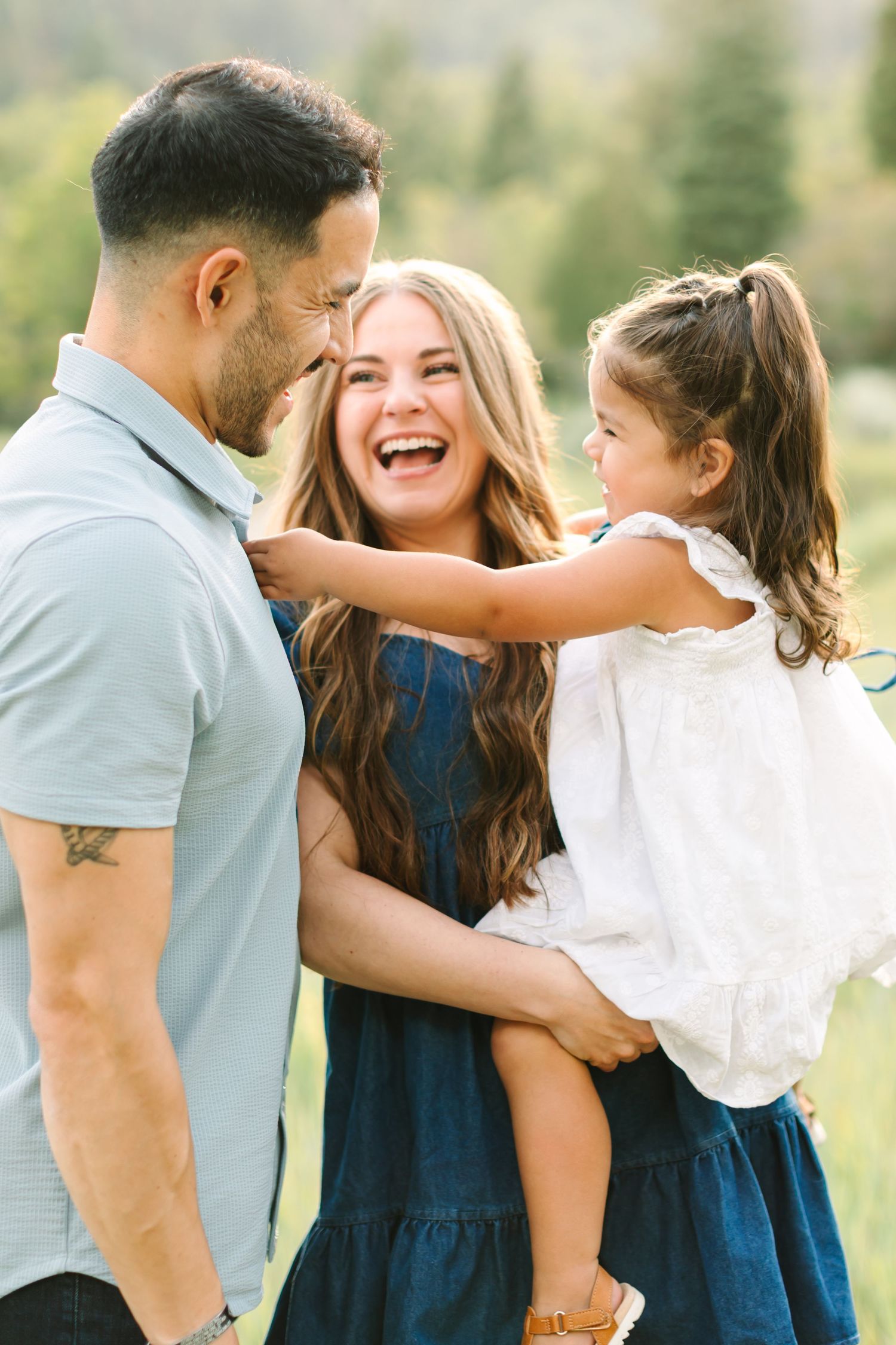 A joyful family of three laughs together outdoors, with parents holding their young daughter in a green field.
