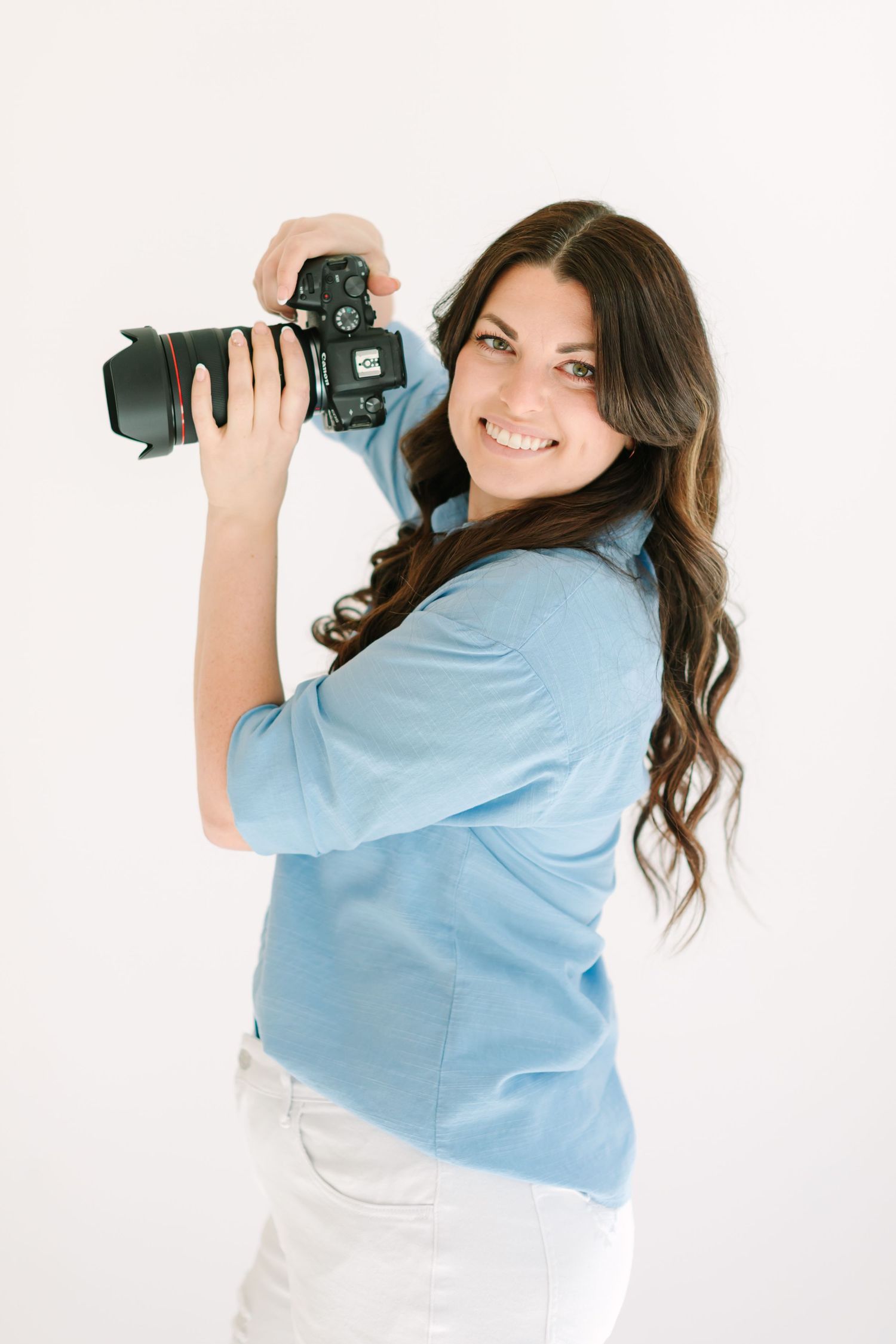 A photographer in a light blue shirt holds a professional DSLR camera against a white background.