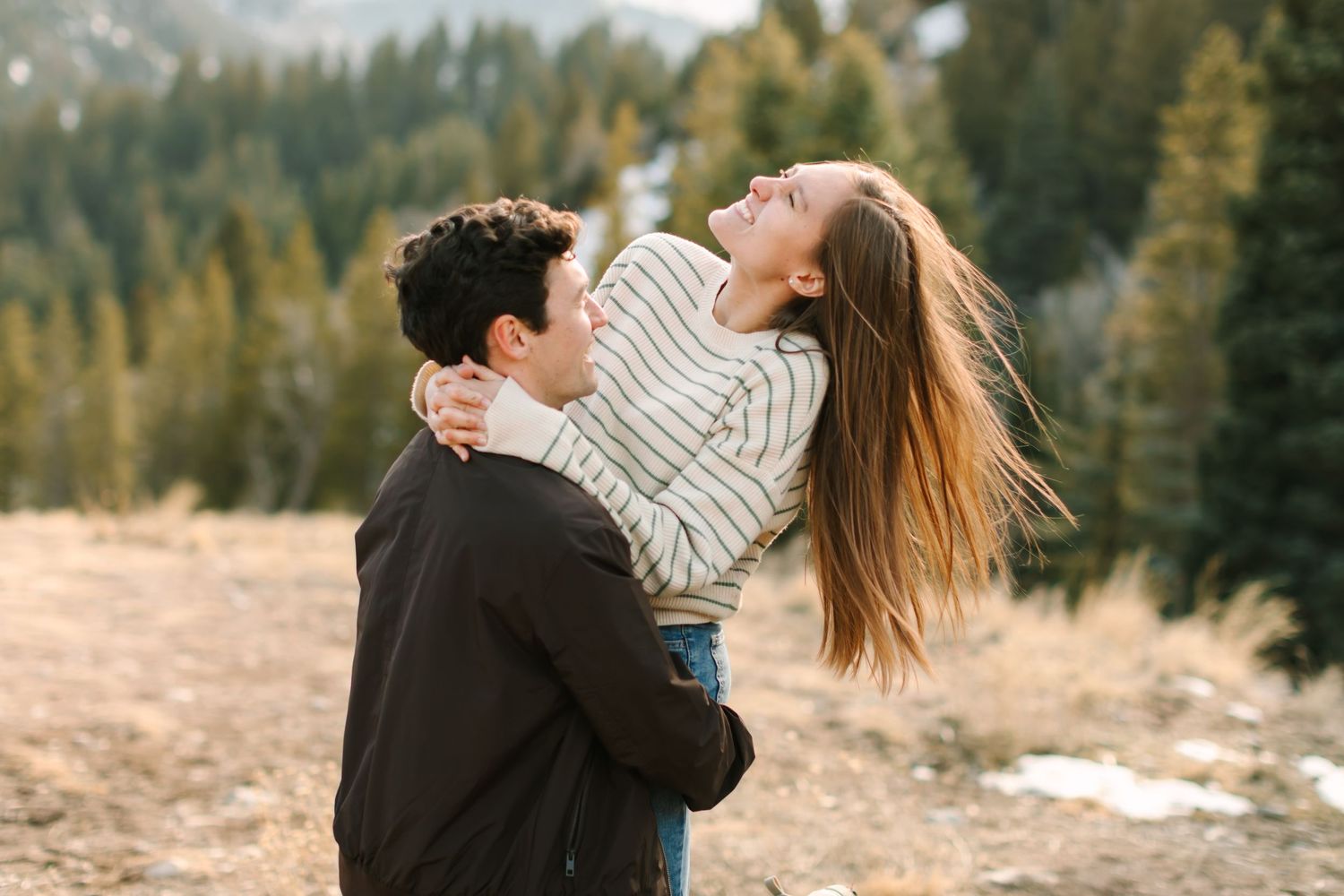 A romantic couple embraces in a snowy mountain landscape surrounded by evergreen trees during a winter photoshoot.