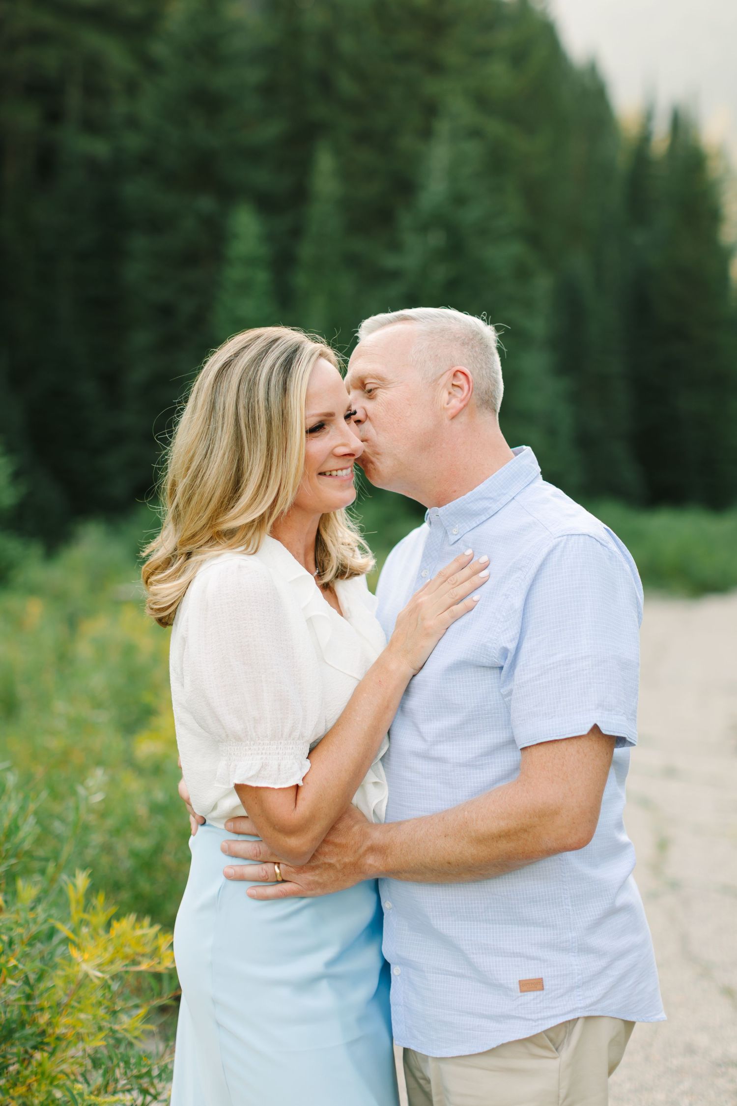A couple embraces outdoors near pine trees, man kissing woman's cheek as she smiles, wearing light blue and white outfits in Utah.
