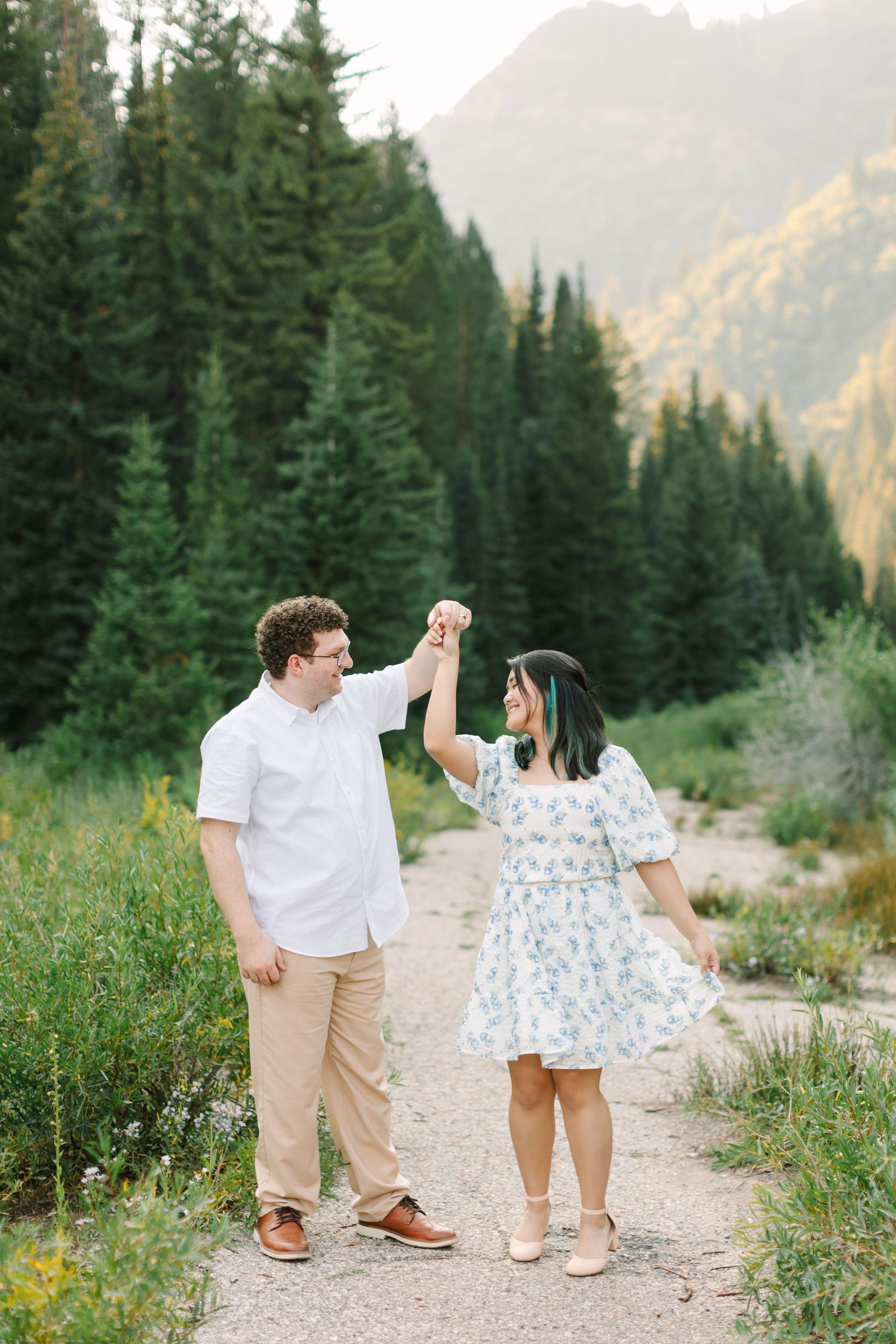 Couple dancing on mountain trail surrounded by pine trees and golden peaks in a romantic outdoor engagement photo in Utah.