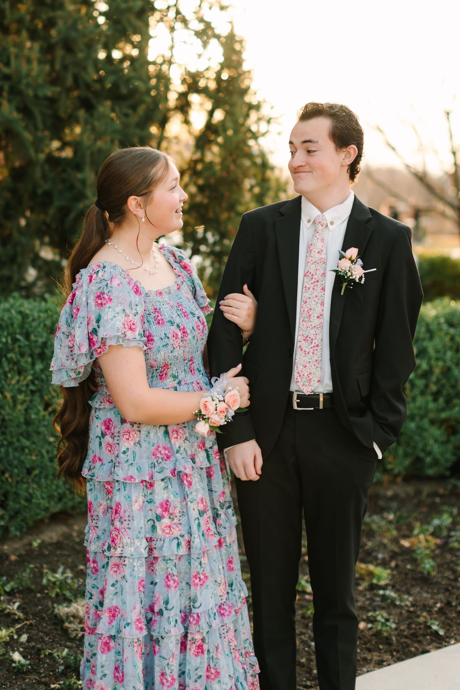 Couple in floral dress and black suit share candid moments during outdoor photo session at sunset.