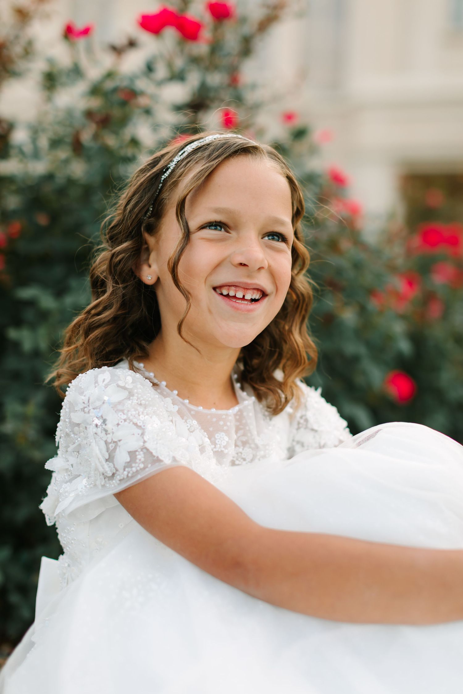 Young girl in white lace dress smiling while standing near red rose bushes in outdoor garden setting.