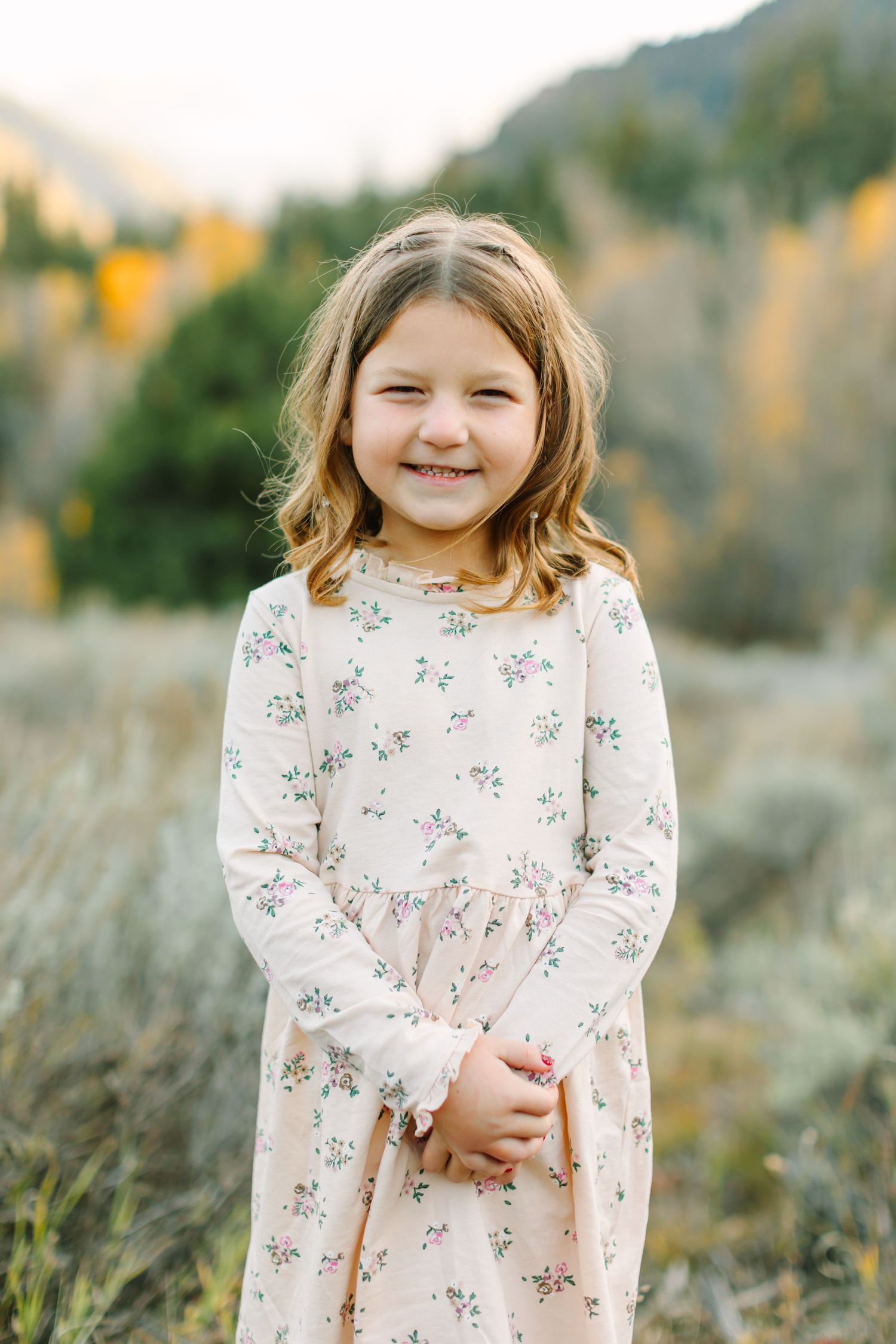 A child wearing a floral dress stands in a field with autumn mountains in the background.