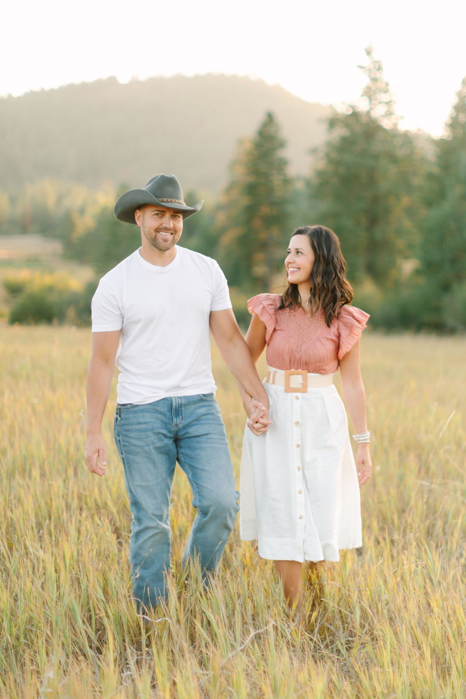 Couple walking hand in hand through a sunlit meadow with mountains in the background.