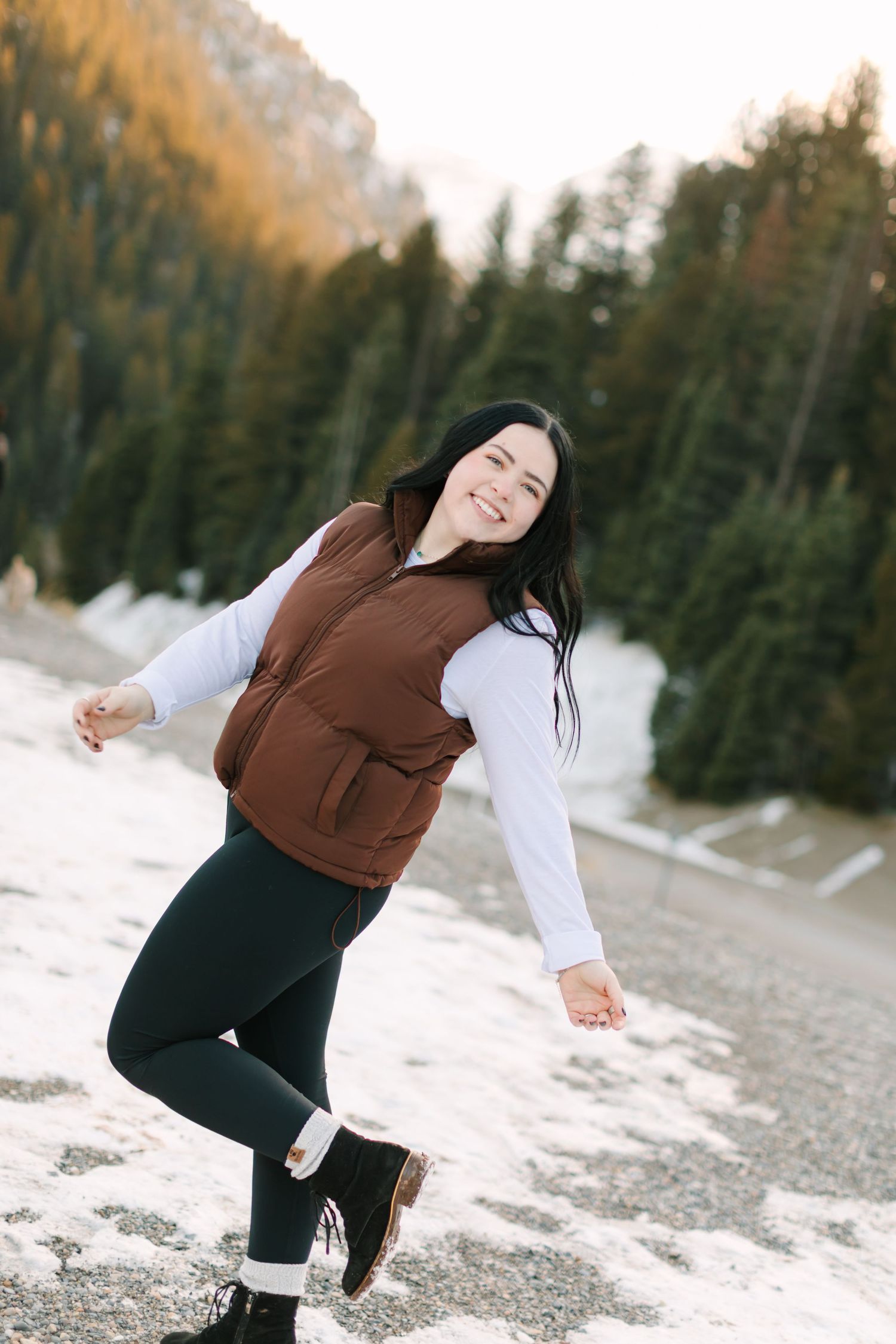 A person in a brown sweater and dark pants poses playfully in a snowy winter landscape with evergreen trees in the background.