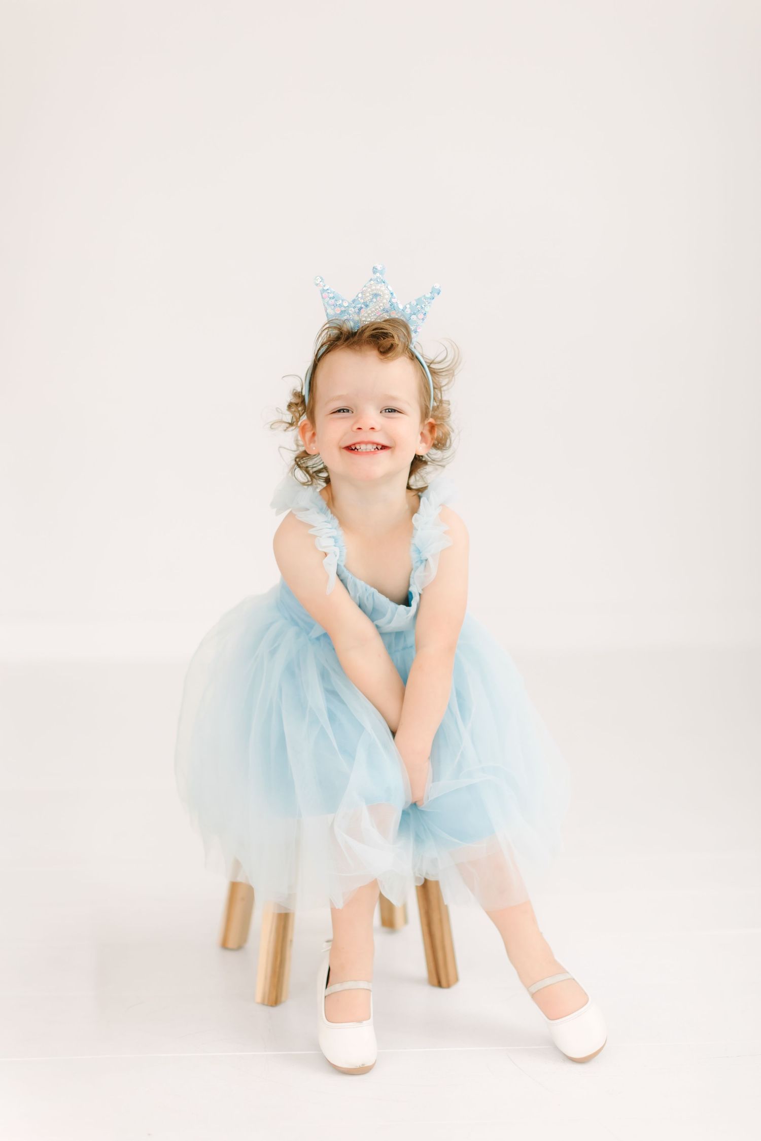 A young girl wearing a blue tulle dress and ballet shoes is smiling and posing on a white background.