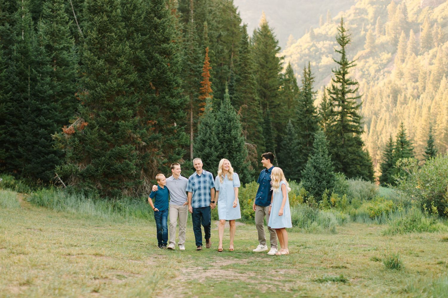 Family of six poses in a lush mountain meadow surrounded by tall pine trees during golden hour in Salt Lake Utah.