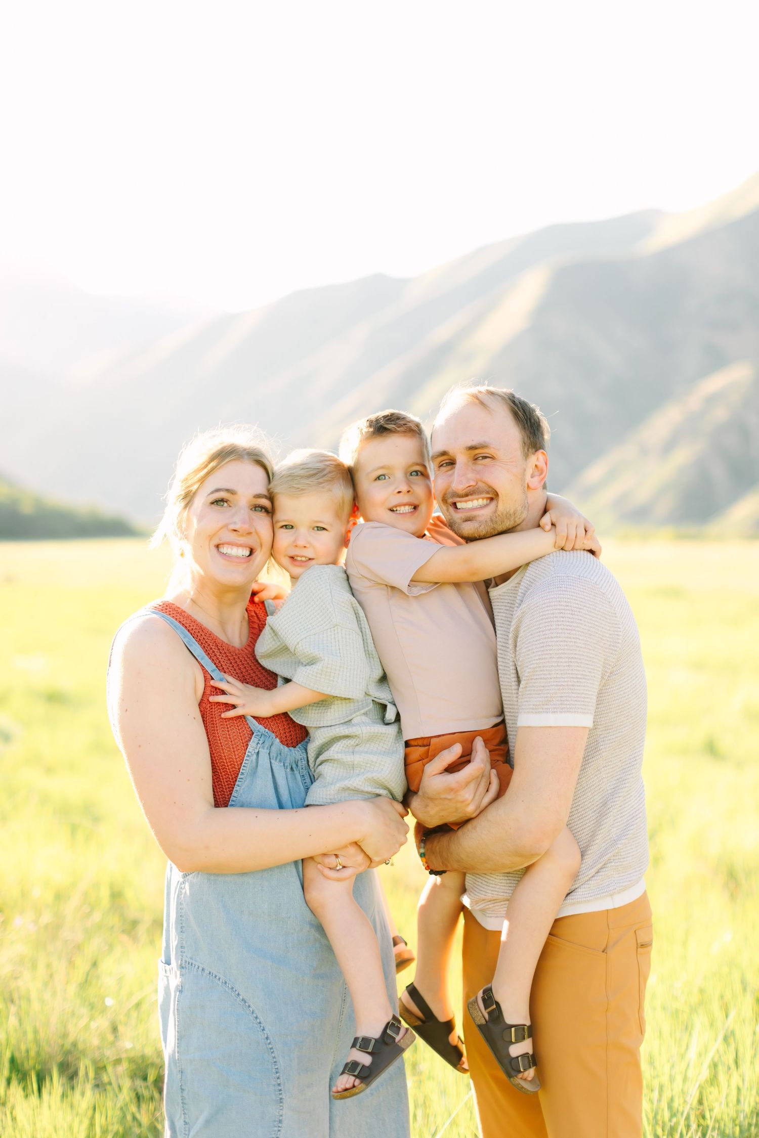 Happy family of four posing in a sunny green field with mountain backdrop, parents holding two young boys.